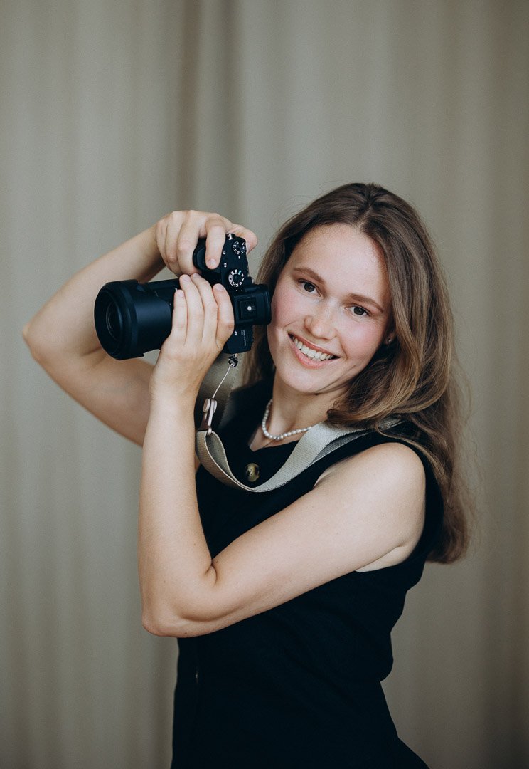 Frau mit braunen Haaren, schwarzen Kleid, Perlenkette, lächelnd, hält Kamera an Kopfhöhe, fotografiert, im Hintergrund eine graue Wand.