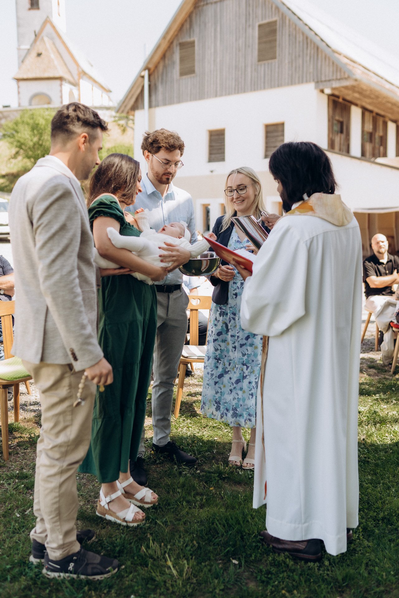 Eine Gruppe von Menschen bei einer Taufe im Freien, mit einem Priester und einem Baby, das getauft wird.