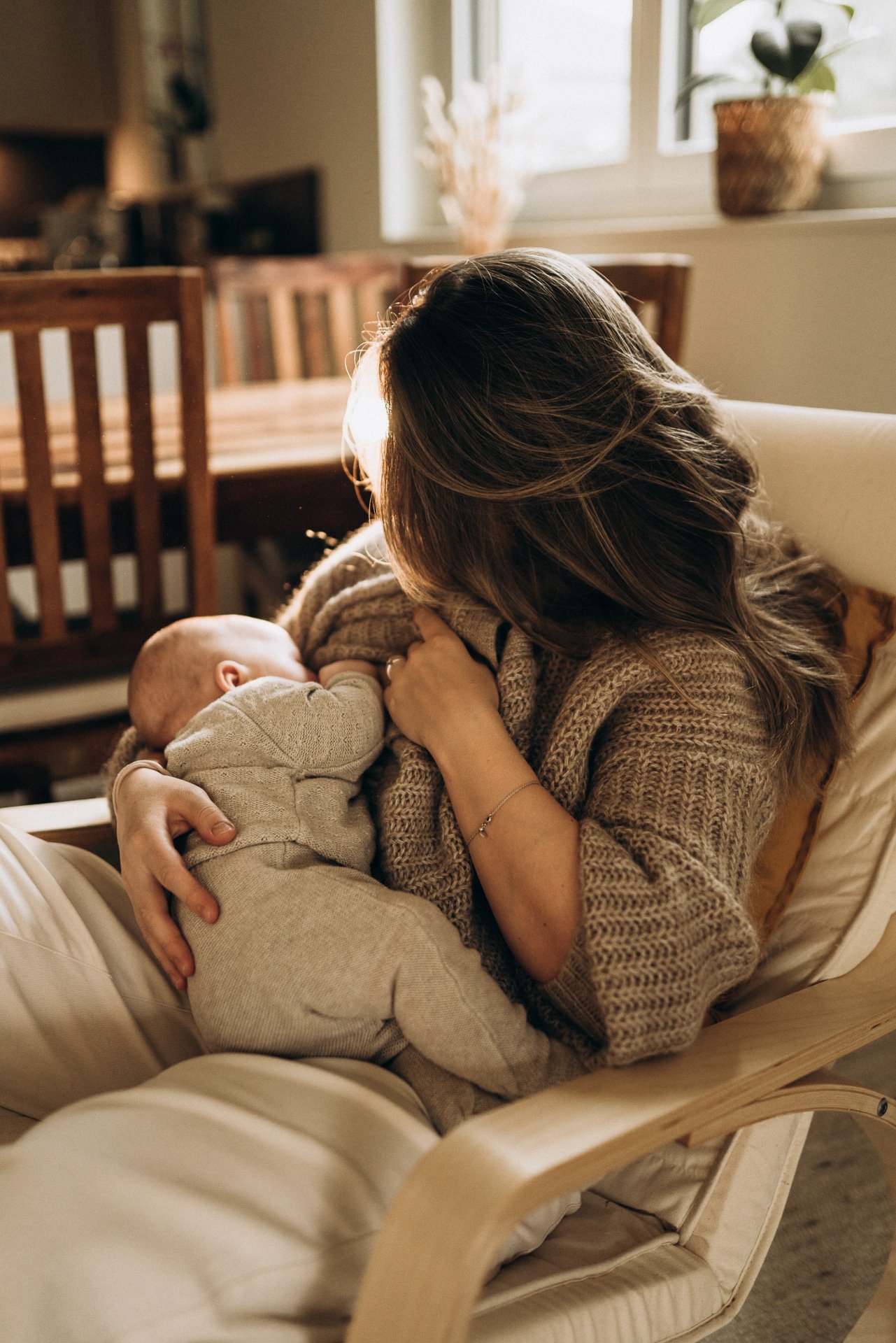 Eine Frau mit braunen Haare stillt ihr Baby in einem gemütlichen Raum, der natürliches Licht durch ein Fenster erhält, mit Pflanzen im Hintergrund und einem Holzlotus-Sessel.