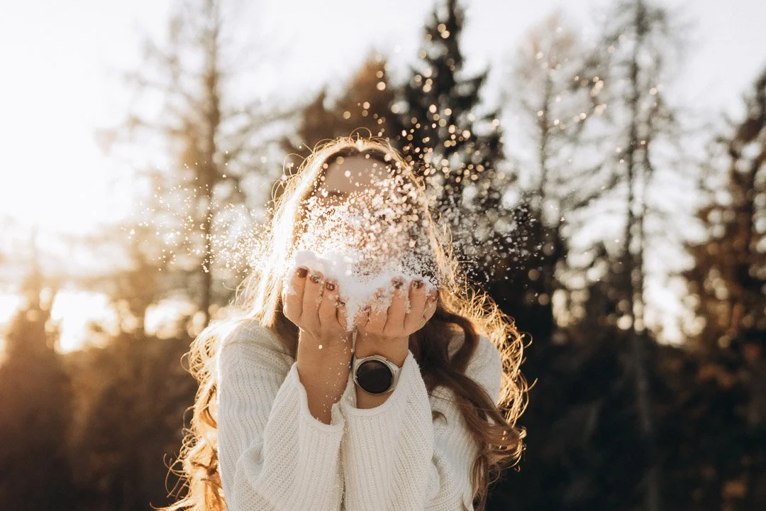 Frau im weißen Pullover wirft Schnee in die Luft vor einem Sonnenuntergang im Wald.
