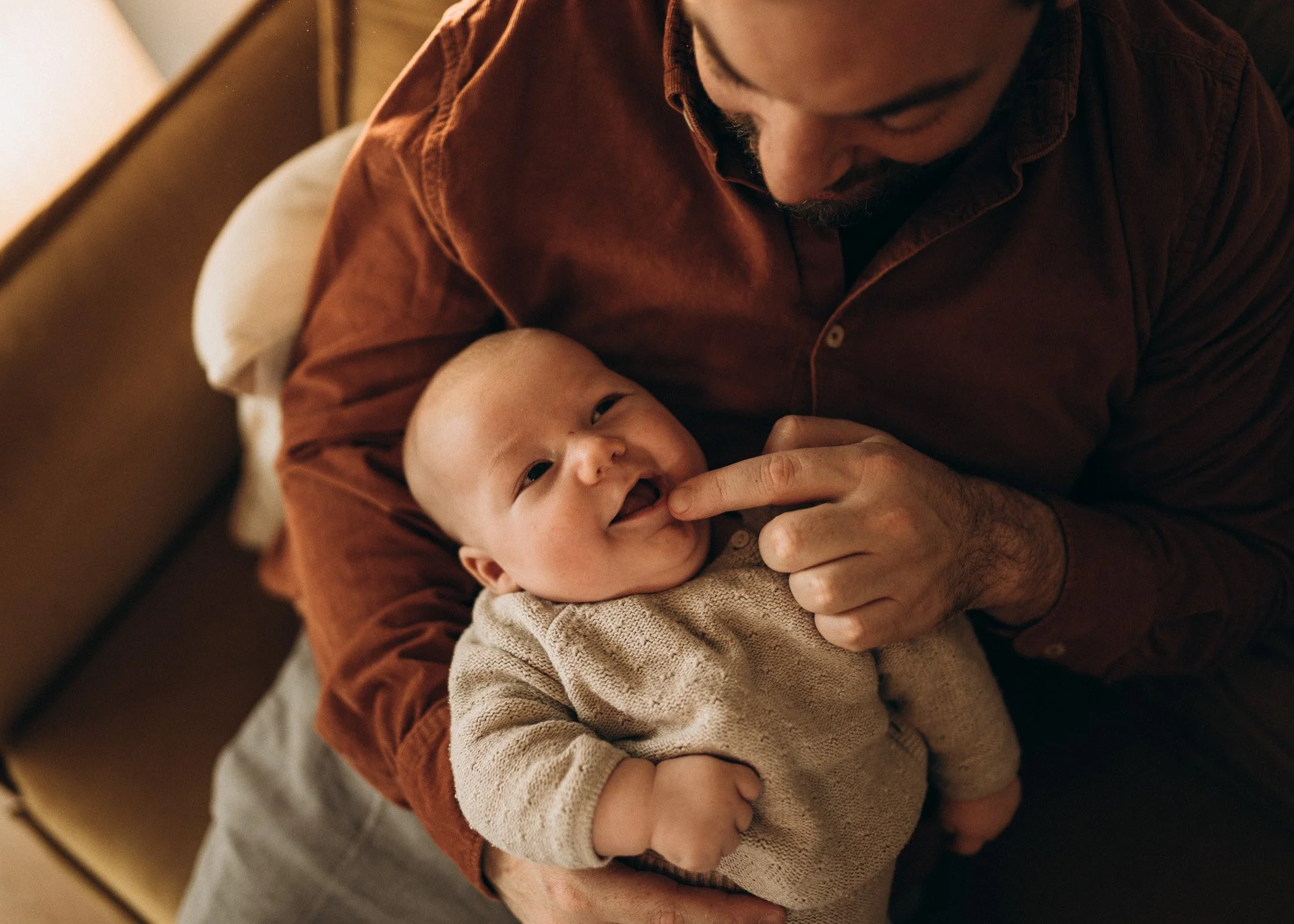 Ein Mann hält ein lächelndes Baby in den Armen, während er es liebevoll berührt. Das Baby trägt einen beige Pullover.