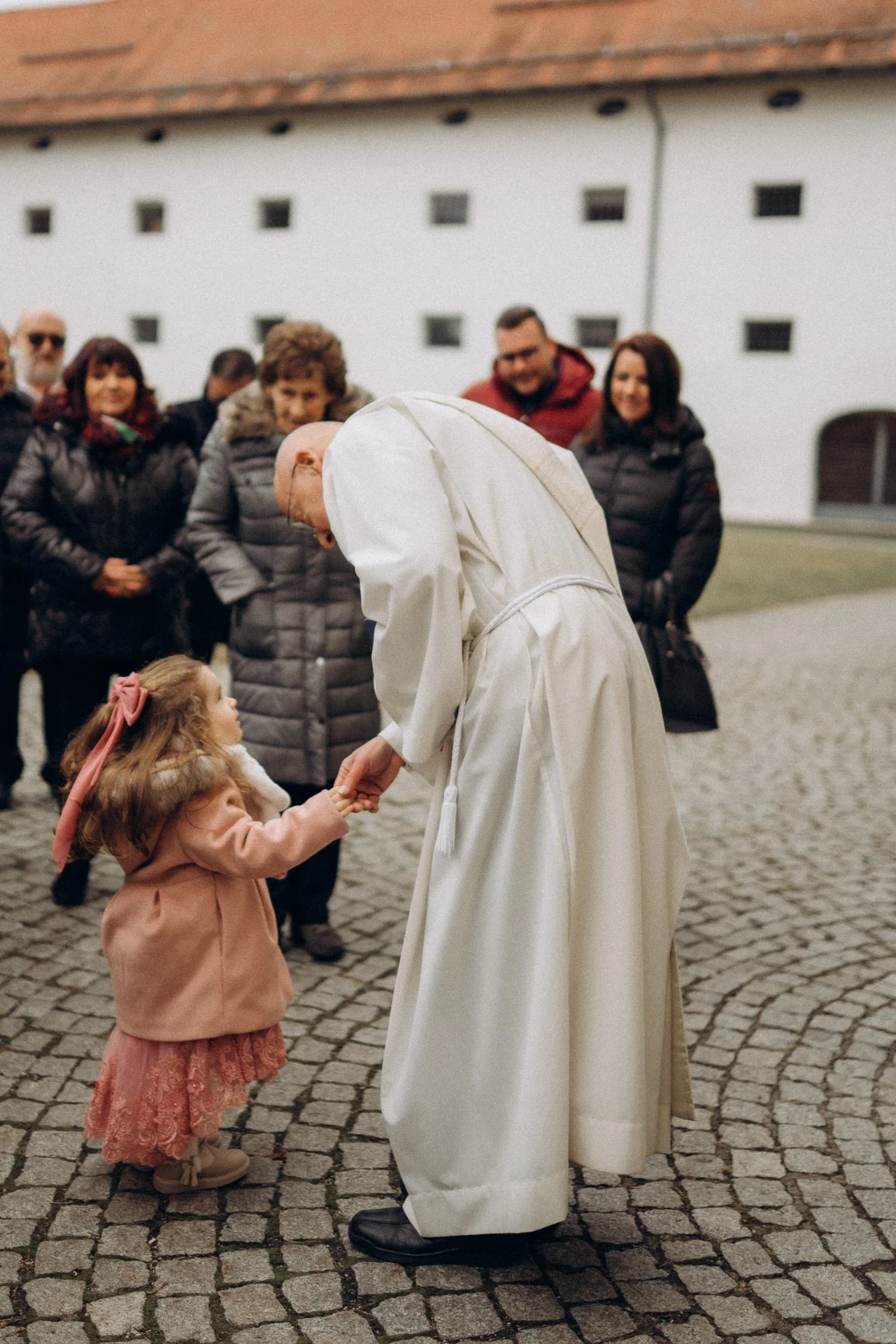 Ein Kardinal in weißem Gewand hält die Hand eines kleinen Mädchens, das ihn anlächelnd ansieht, während eine Gruppe von Menschen im Hintergrund zuschaut.