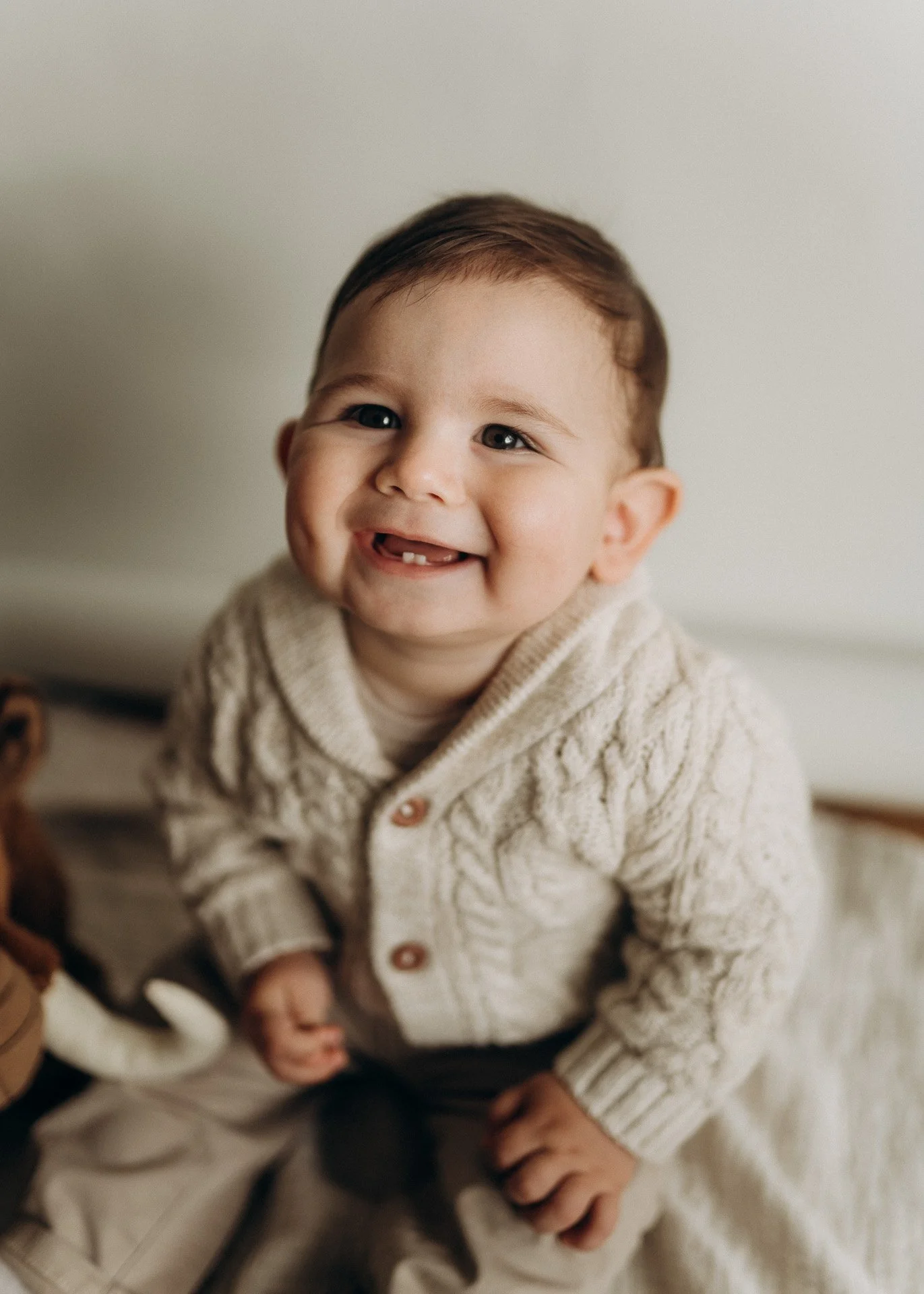 Ein glückliches Baby mit braunen Haaren und blauen Augen trägt einen cremefarbenen Strickpullover mit Knöpfen.