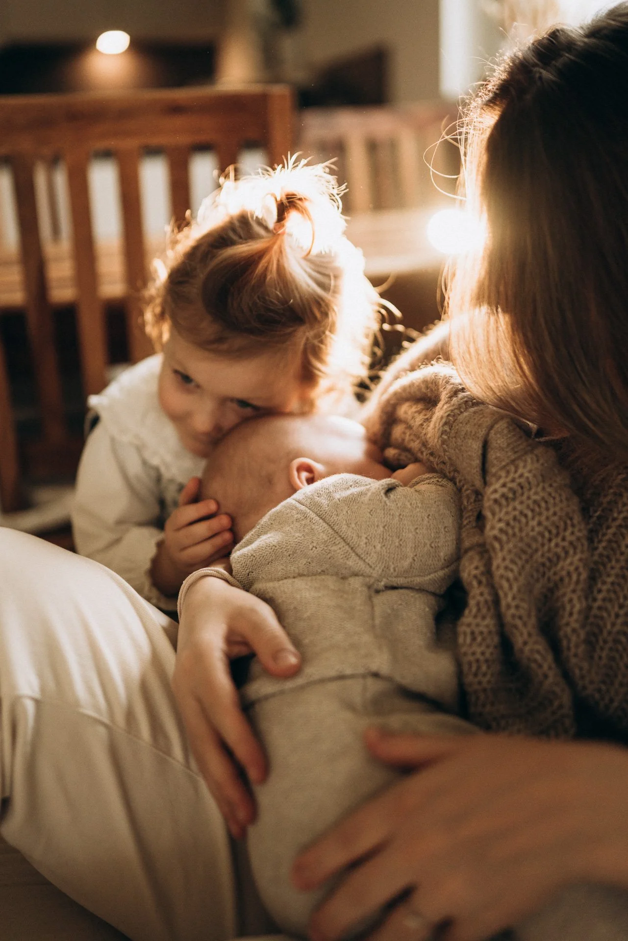 Eine Frau und zwei Kinder, eines davon ein Baby, in einer warmen, gemütlichen Umgebung, vermutlich beim Kuscheln oder Kosen. Die Sonne scheint durch ein Fenster, was eine warme Atmosphäre schafft.