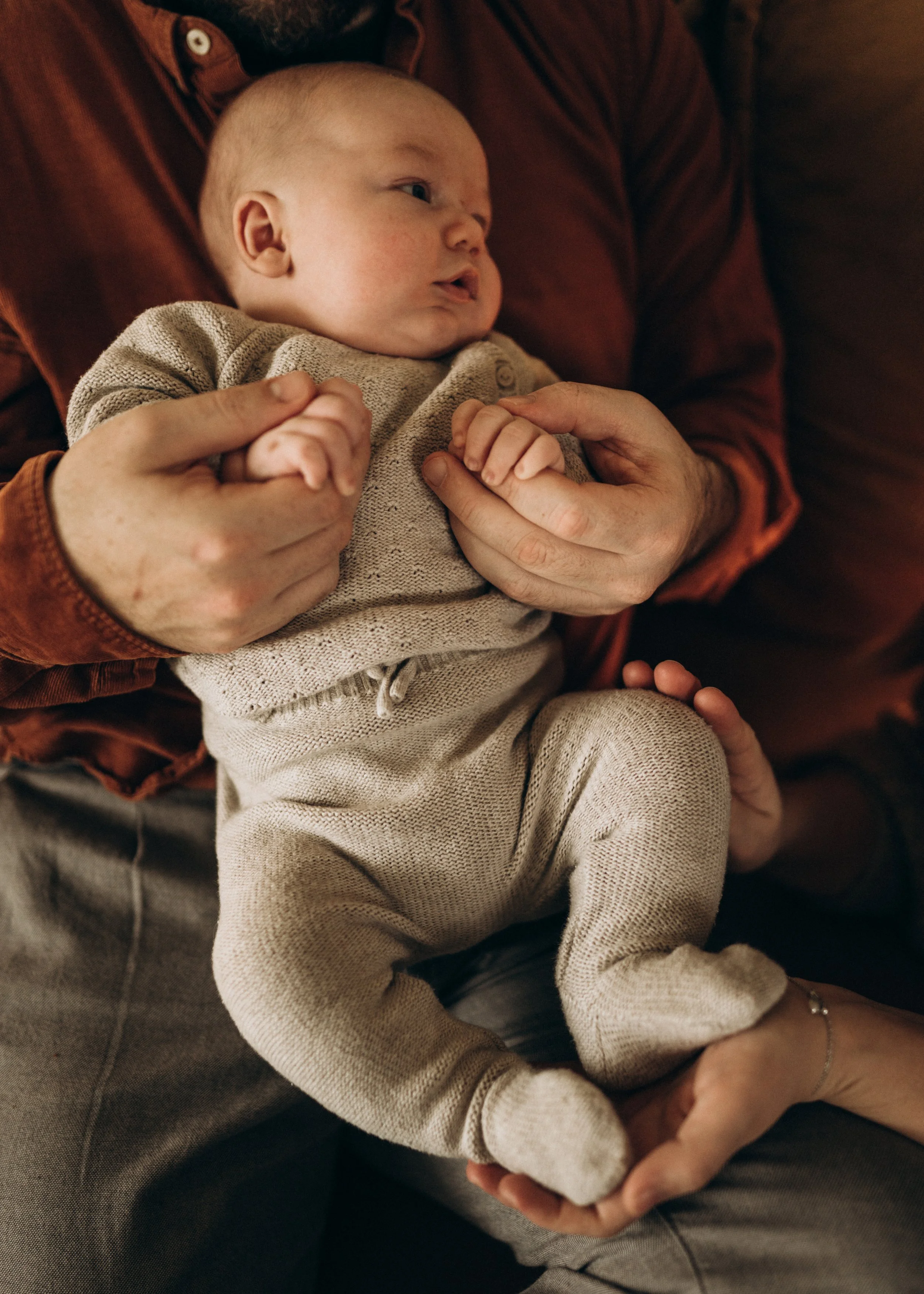 Ein Baby liegt auf einer Person, die ihn liebevoll hält, beide sitzen auf einem Sofa, neutral gekleidet, in einer warmen, einladenden Umgebung.