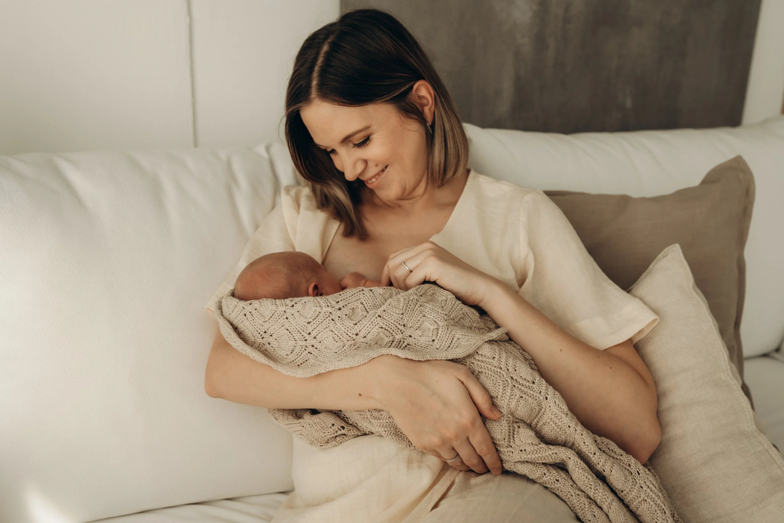 Eine Frau hält ein neugeborenes Baby in ihren Armen auf einem Bett. Sie lächelt liebevoll und schaut auf das Baby, das in eine gestrickte Decke eingewickelt ist.