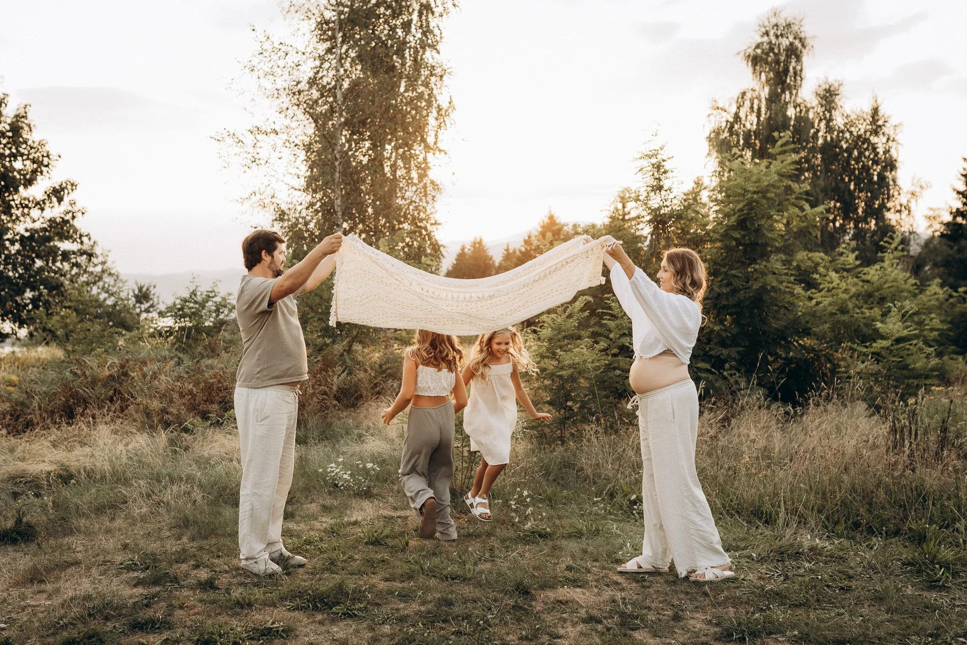Eine Familie mit zwei Kindern feiert im Freien bei Sonnenuntergang, Eltern halten eine Decke, Kinder tanzen fröhlich.