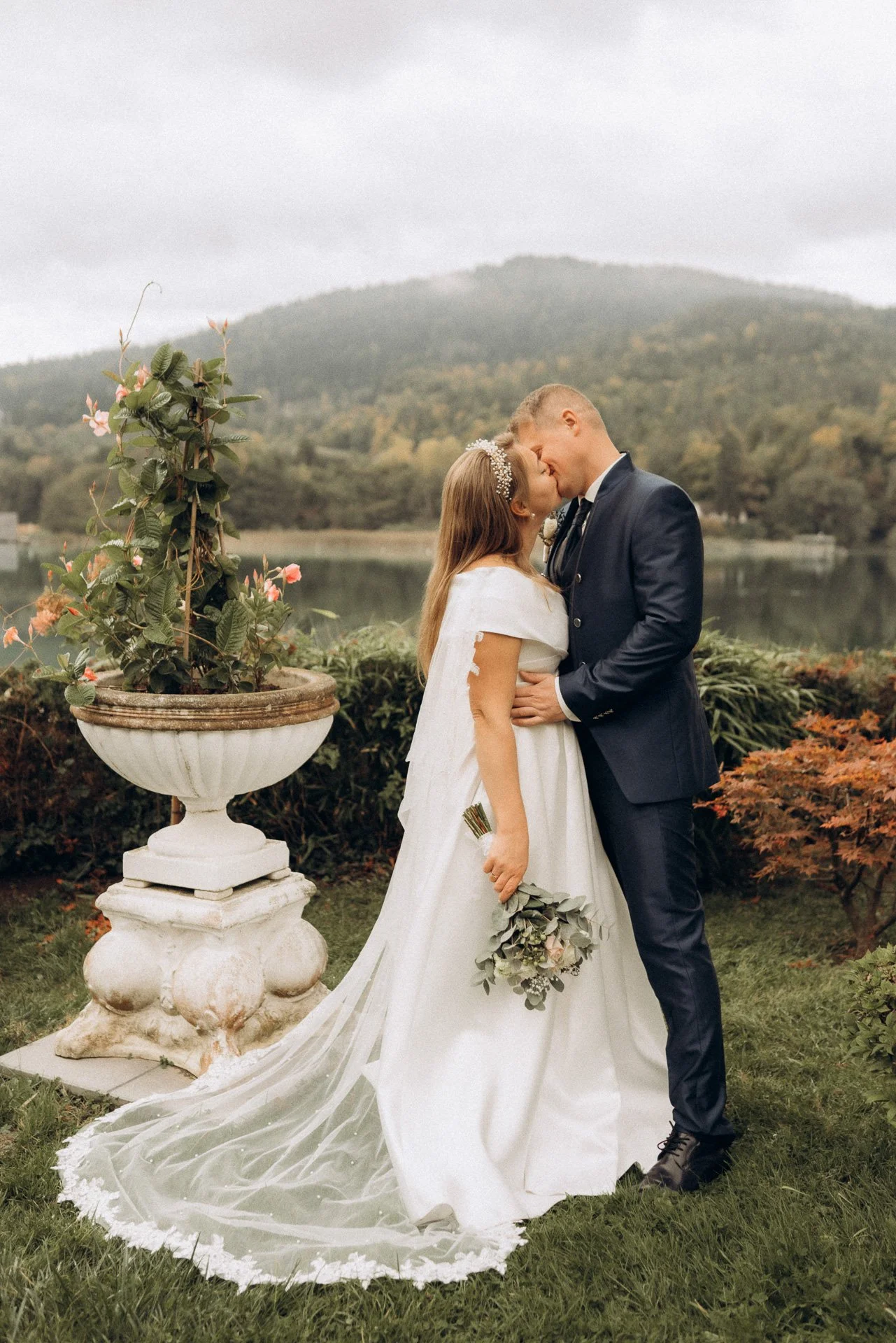 Ein Brautpaar küsst sich bei einer Hochzeit im Freien, mit einem Fluss und Hügel im Hintergrund, umgeben von Herbstblättern und einer großen Blumenvase mit Blumen.