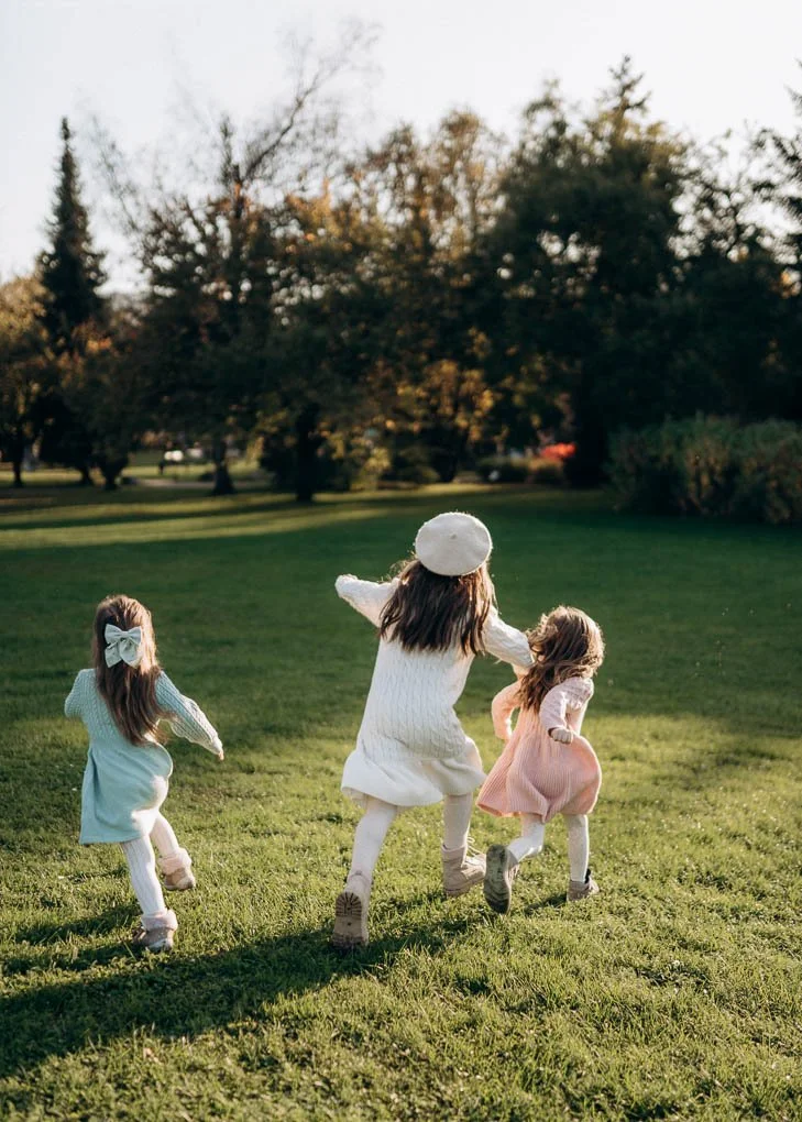 Drei kleine Mädchen laufen lachend über eine grüne Wiese im Park, aufgenommen während eines Familienshootings im warmen Licht des Sonnenuntergangs in Klagenfurt, Kärnten.