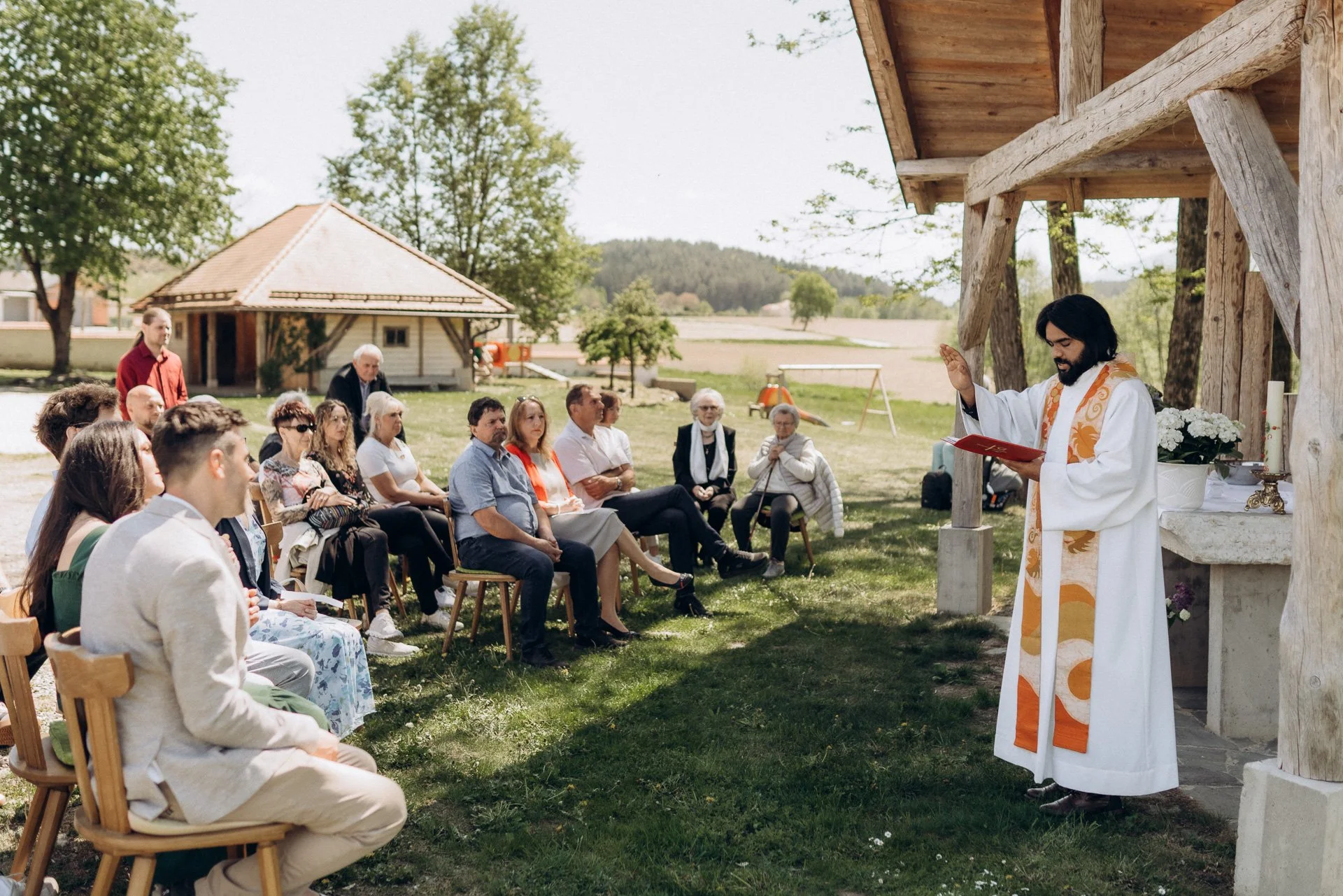 Ein Priester officiert eine Zeremonie im Freien vor einer Gruppe von Menschen, die im Garten sitzen.
