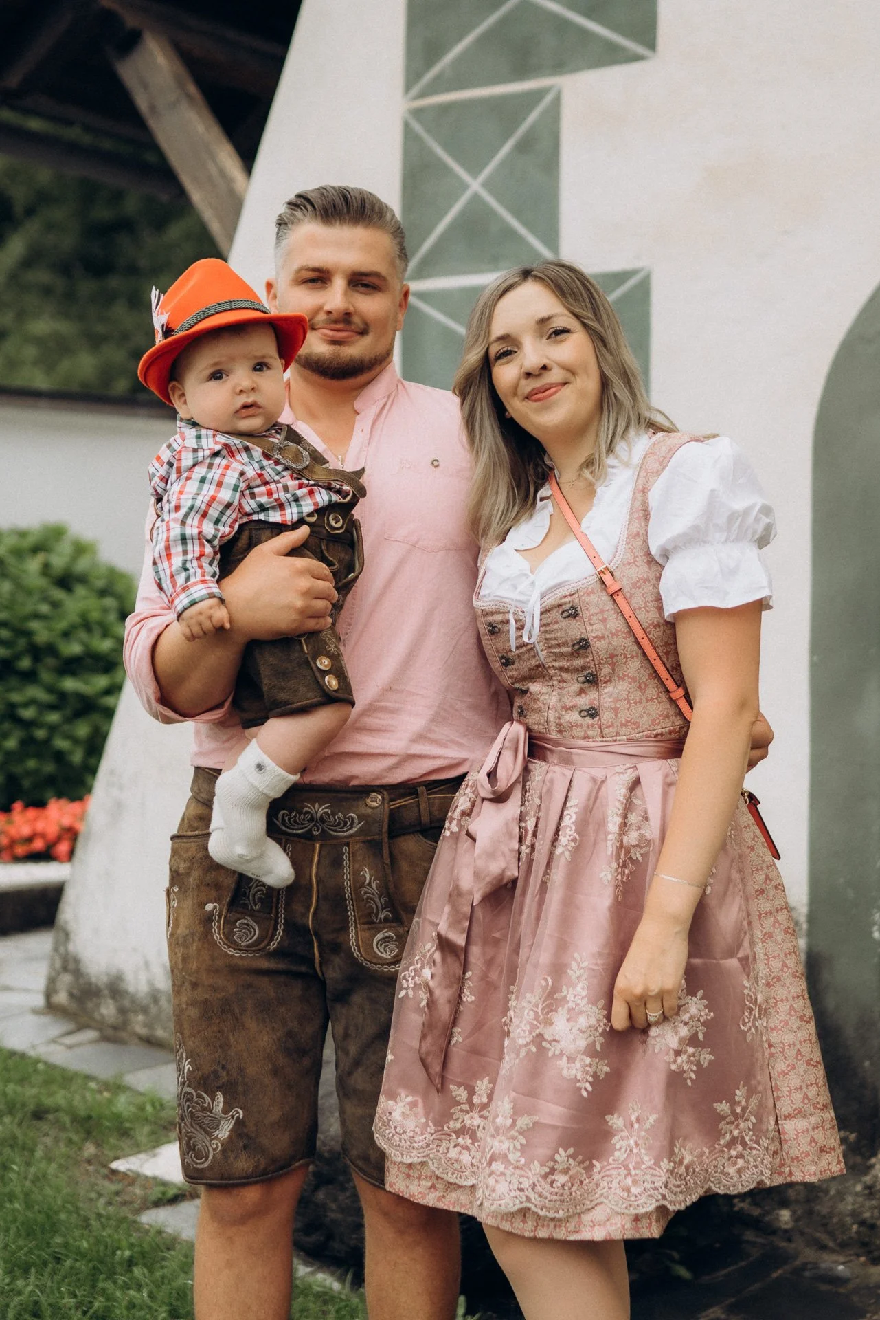 Familie bei einem traditionellen Fest, der kleine Junge trägt einen roten Hut mit Feder, die Erwachsenen tragen Trachten, im Hintergrund ein weißes Gebäude.