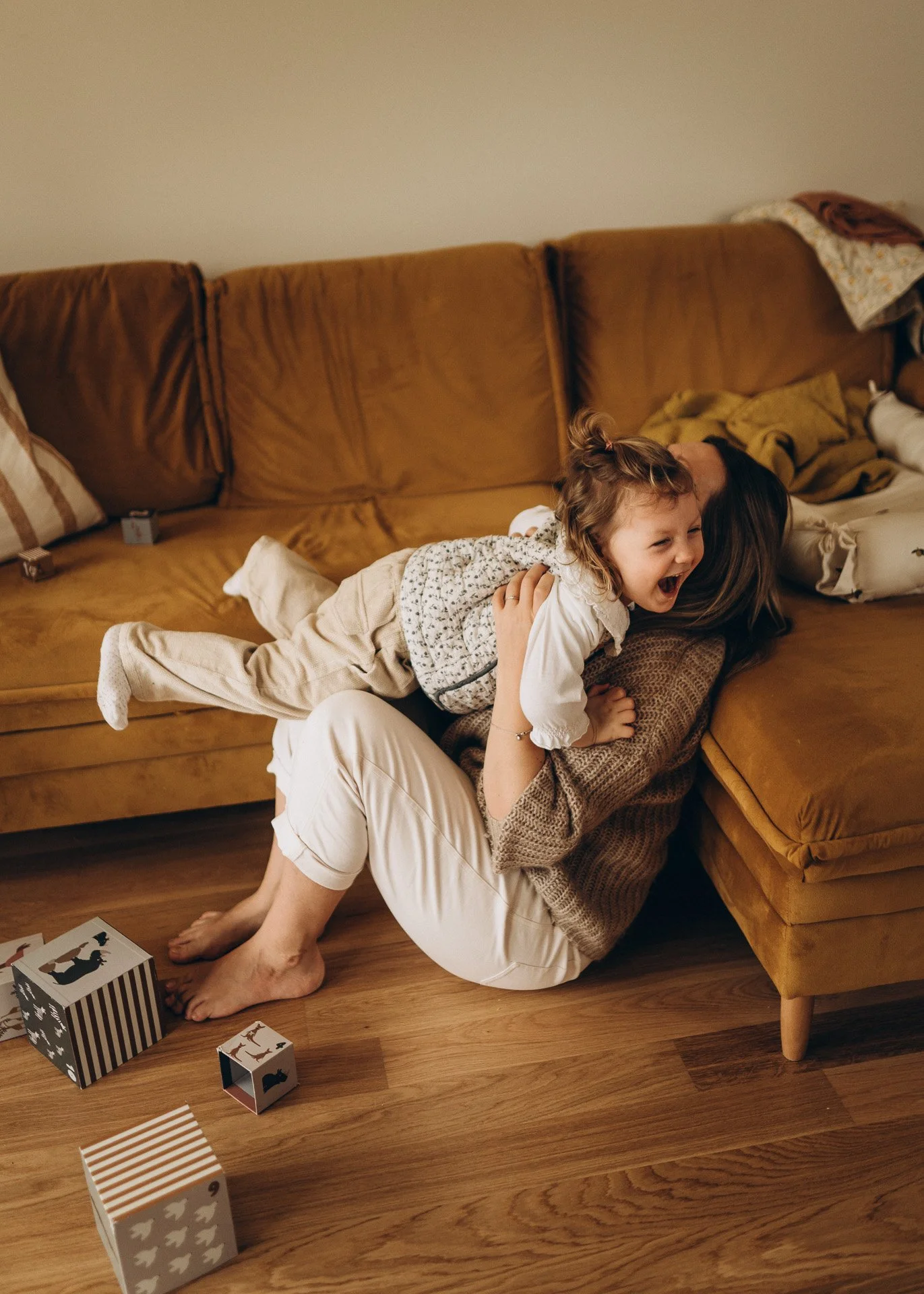 Zwei Kinder und eine erwachsene Person liegen lachend auf einem goldbraunen Sofa. Es gibt Spielzeugwürfel auf dem Holzboden, und das Zimmer ist gemütlich eingerichtet.