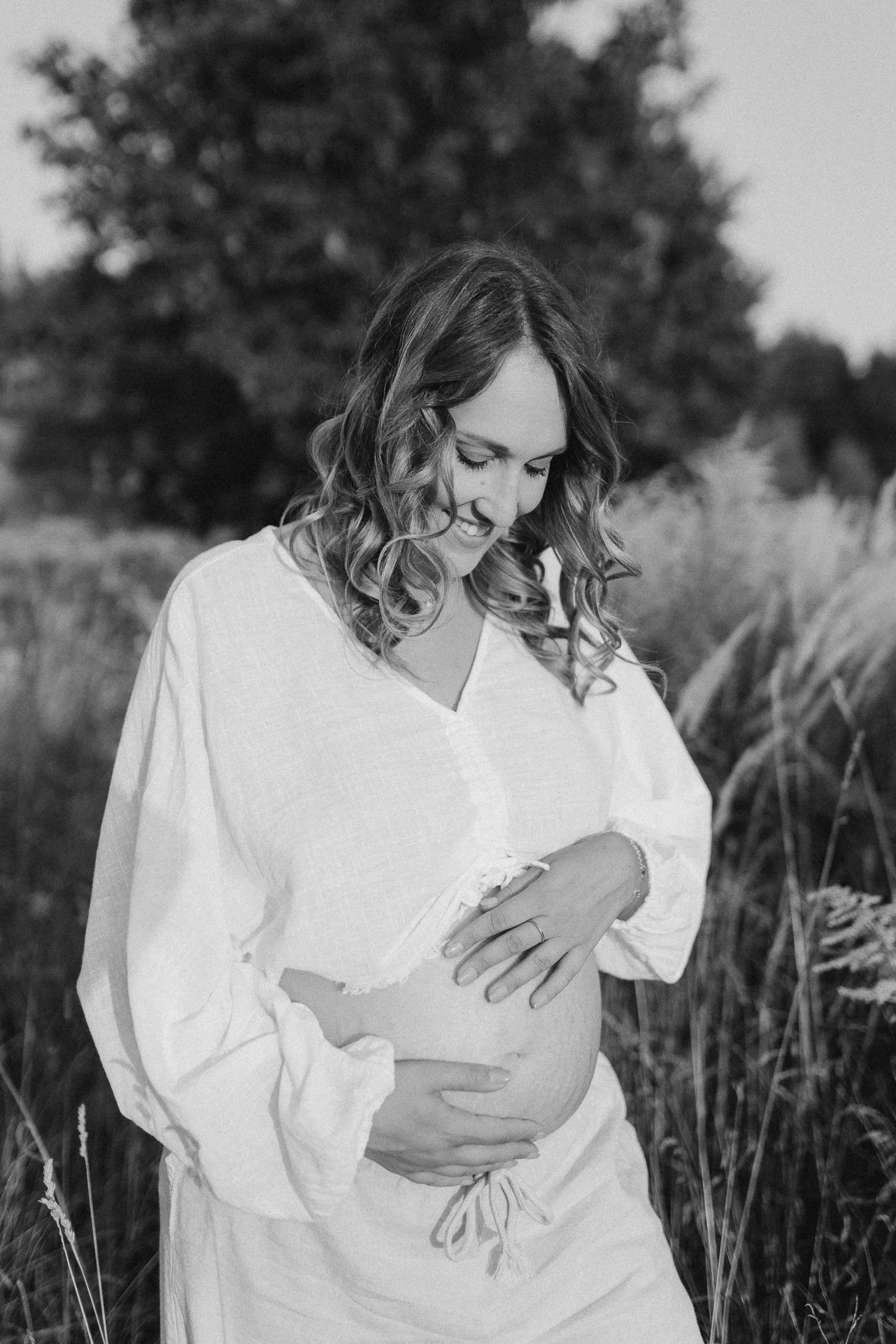 Schwangere Frau in einem weißen Kleid im Freien, die ihren Bauch hält, in Schwarz-Weiß-Foto aufgenommen.