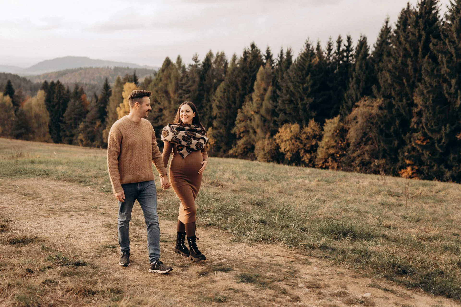 Ein Paar geht Hand in Hand durch ein Herbstwaldgebiet, die Frau ist schwanger und trägt Schal und schwarze Stiefel, der Mann trägt einen braunen Pullover und Jeans.