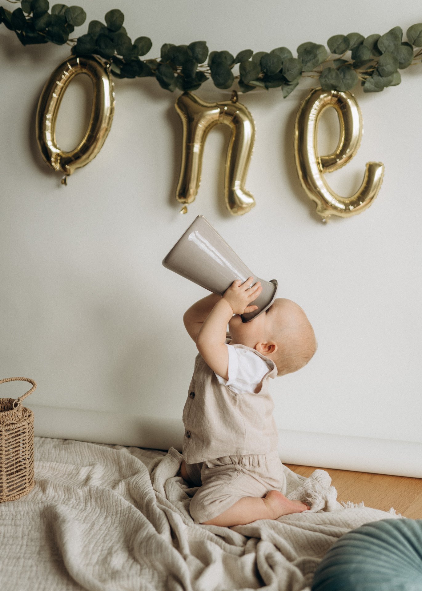 Ein Baby sitzt auf einem weichen, cremefarbenen Tuch am Boden und spielt mit einer großen, grauen Kissen. Es hält einen grauen Luftballon, der Schaumstoff ist, und hebt ihn über den Kopf. Im Hintergrund sind goldene Ballons in Form von Buchstaben, di