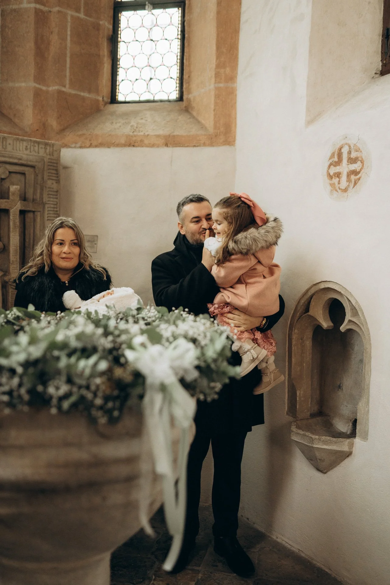 Familie bei einer Taufe in einer alten Kirche, mit Blumen im Vordergrund und einem steinernen Altar.