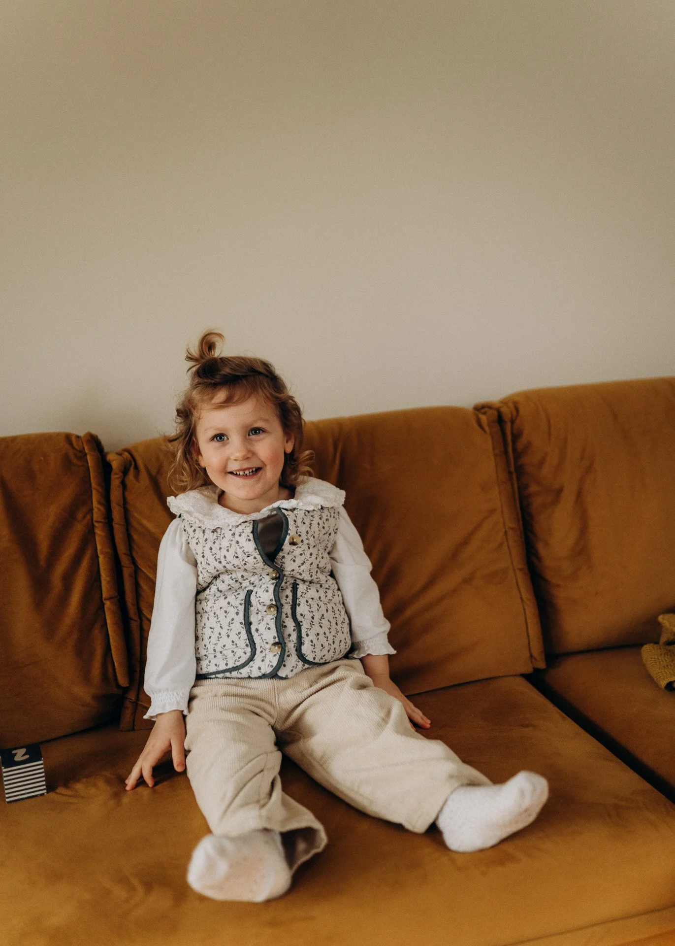 Ein lachendes Mädchen sitzt auf einer braunen Couch vor einer beige Wand. Sie trägt eine weiße Bluse, eine bestickte Weste, beige Hosen und weiße Socken.