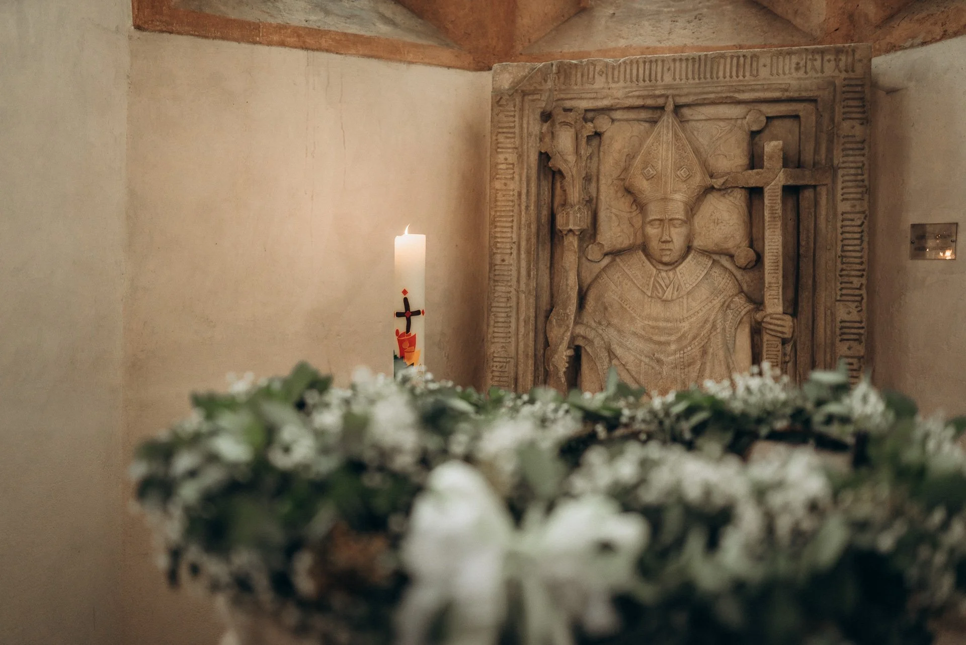 Ein geschnitztes Relief eines Heiligen, eine brennende Kerze mit Kreuz und Blumen im Vordergrund.