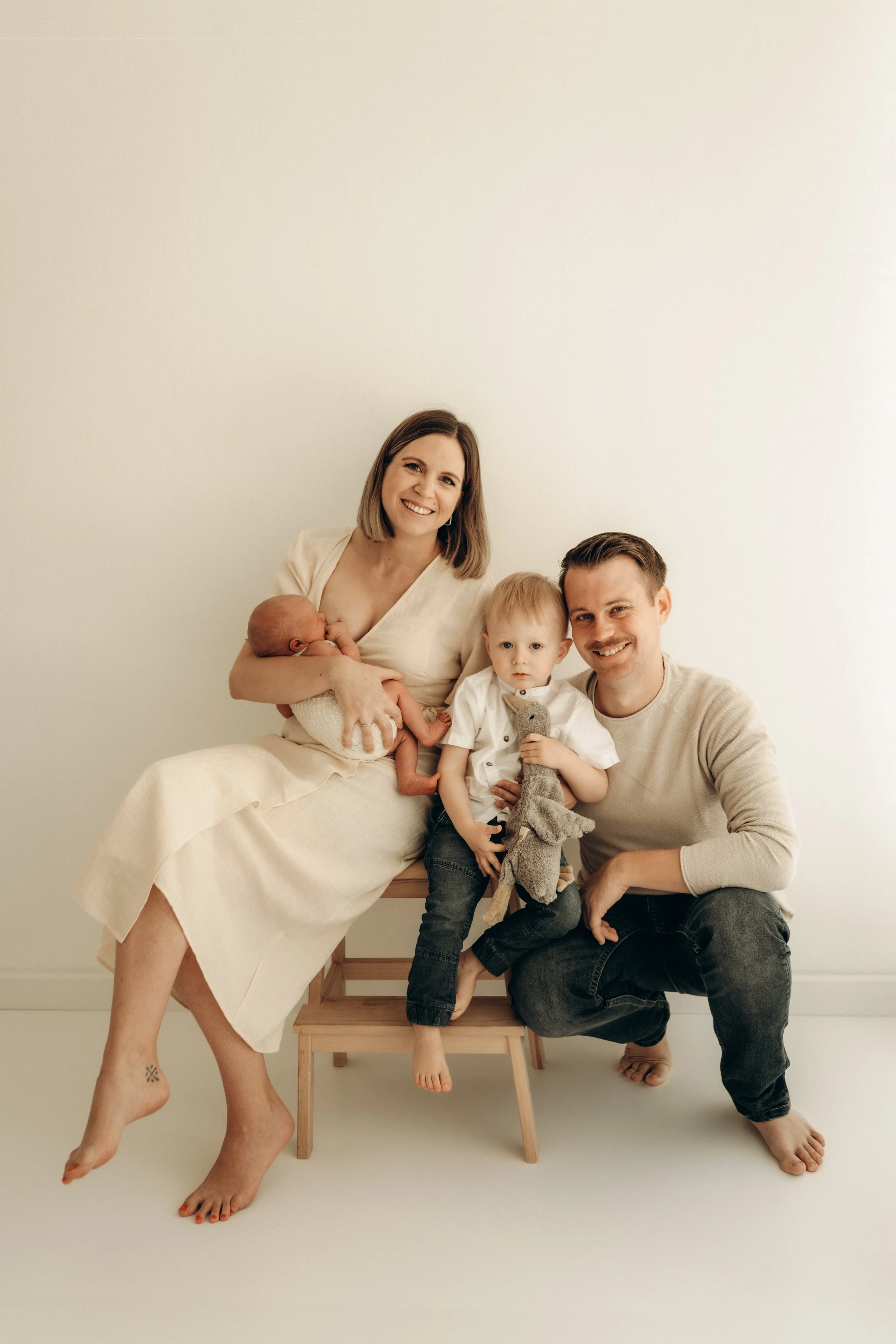 Familie mit zwei kleinen Kindern sitzt vor einem weißen Hintergrund, die Mutter stillt das Baby, während der Vater und das ältere Kind neben ihr sitzen, alle lachen und schauen in die Kamera.