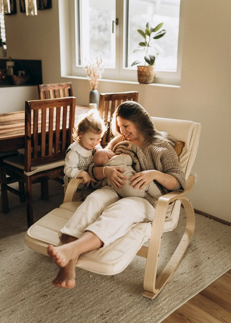 Eine Mutter sitzt auf einem Schaukelstuhl, hält ein Baby im Arm und hat ein Lächeln im Gesicht, während ihre Tochter neben ihr das Baby betrachtet, in einem hell erleuchteten Wohnzimmer.