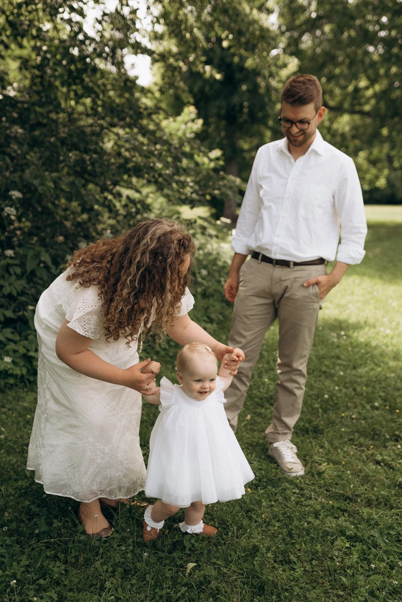 Eine Frau mit lockigem Haar hält ein Baby, während ein Mann im weißen Hemd und Brille lächelnd zuschaut, im Grünen.