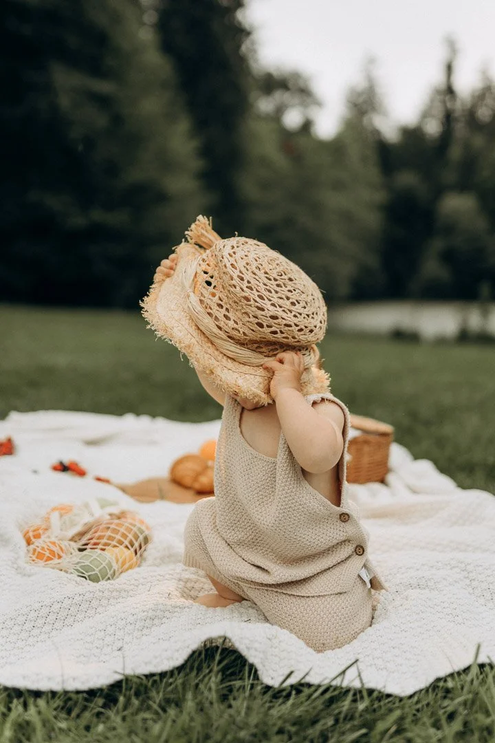 Kleines Kind mit Strohhut und beigem Strickkleid, sitzend auf einer Picknickdecke im Park während eines sommerlichen Familienshootings in Klagenfurt, Kärnten.
