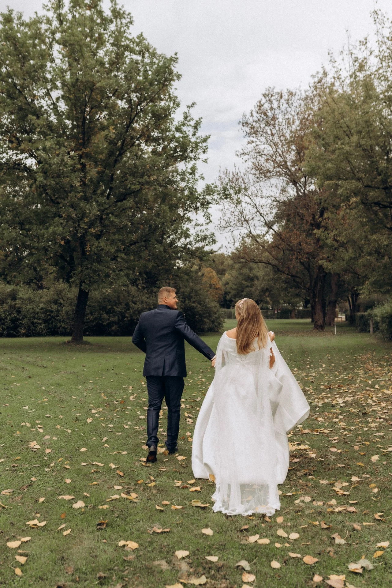 Ein Hochzeitspaar, ein Mann in Anzug und eine Frau in einem weißen Brautkleid, läuft Hand in Hand durch einen parkartigen Garten mit Bäumen und fallenden Blättern.