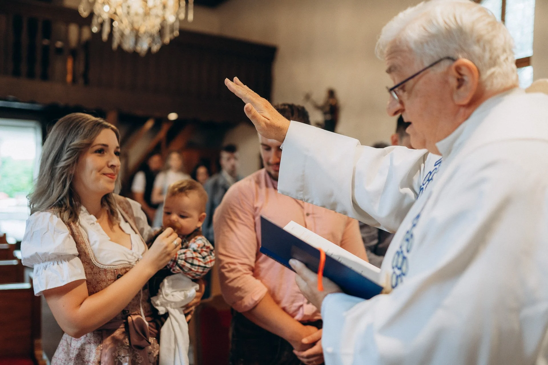 Ein Priester segnet eine Familie bei einer Zeremonie in einer Kirche, die Mutter hält ein Baby, während andere Familienmitglieder zuschauen.