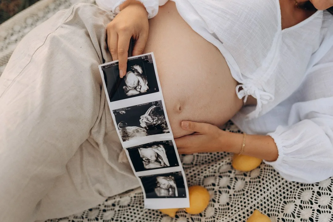 Eine schwangere Frau hält Ultraschallbilder ihres Babys vor den Bauch, während sie auf einem Tisch mit einem eleganten, gelochten Tischtuch sitzt, umgeben von Zitronen.