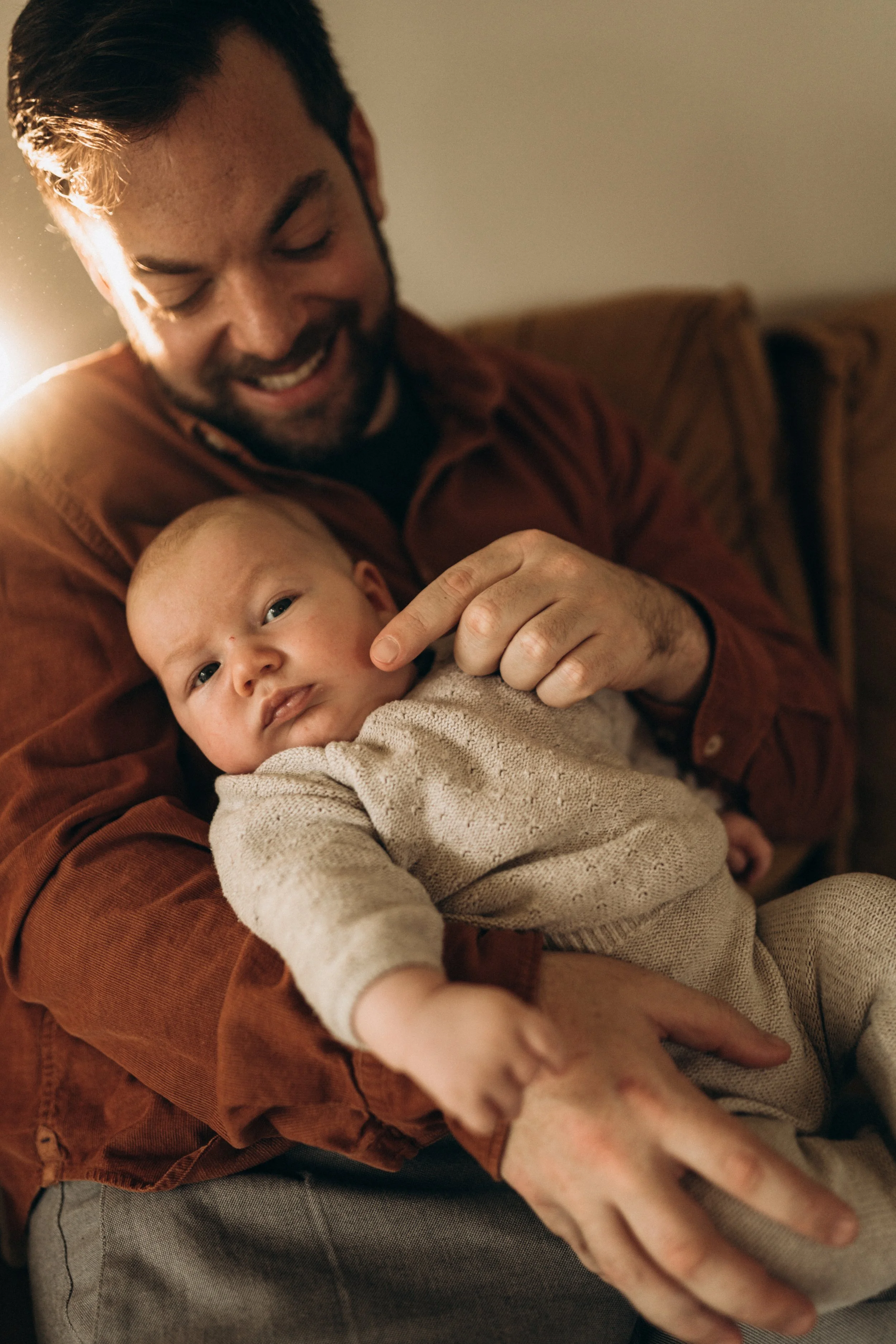 Ein Mann mit dunklem Haar und Bart hält ein kleines Baby in den Armen, beide lachen. Das Baby trägt einen beige Pullover.