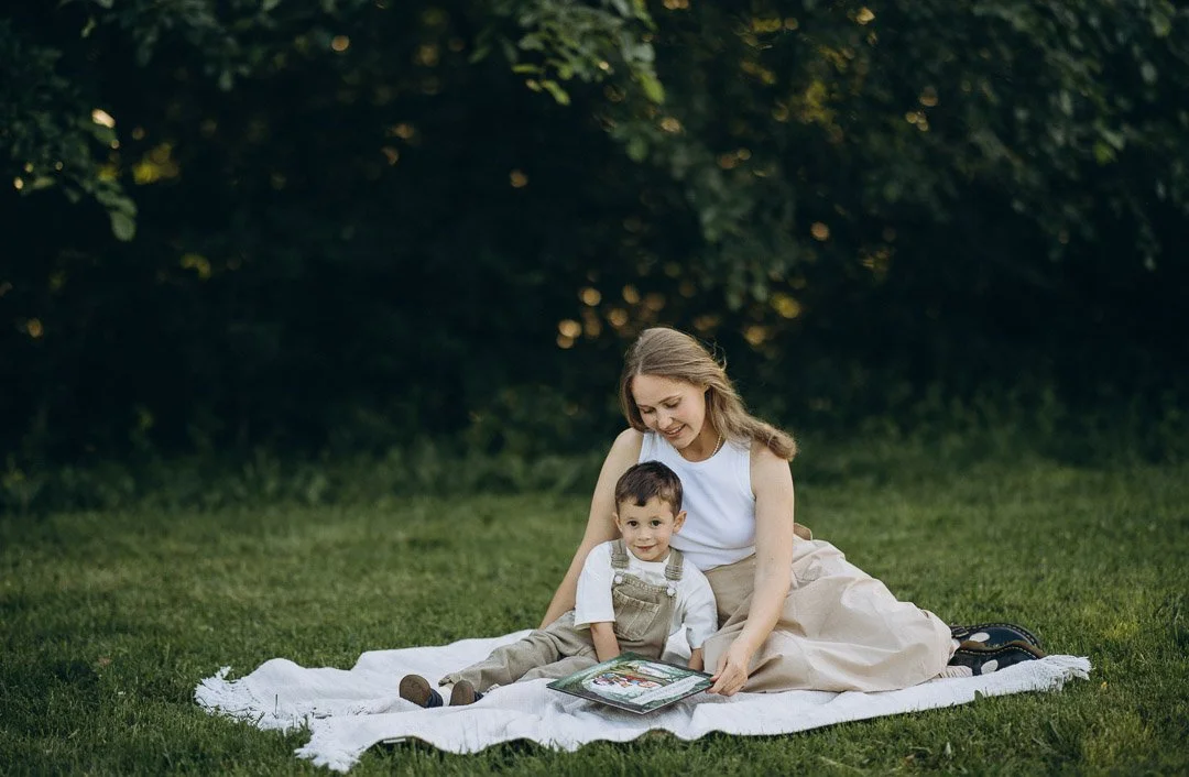 Eine junge Frau liest einem kleinen Jungen auf einer Picknickdecke im Park vor.