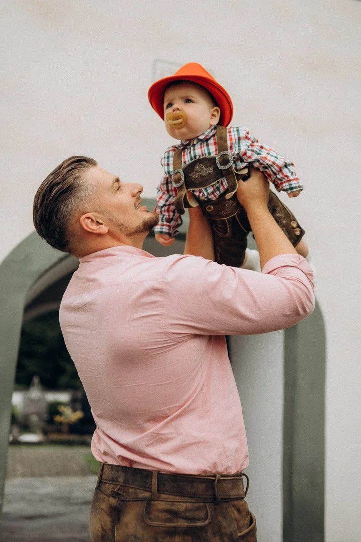 Ein Mann hebt ein Baby mit traditioneller bayerischer Kleidung und Hut hoch, das ein Schnuller im Mund hat.