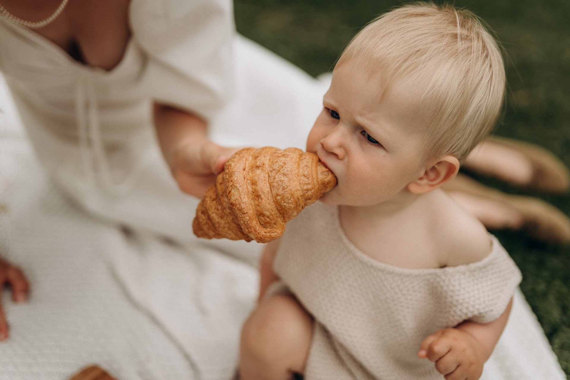 Ein Kind beißt in eine Croissant, gehalten von einer Person, vermutlich die Mutter, im Freien. Das Kind trägt ein hellbeiges Oberteil und hat blondes Haar, die Umgebung ist grün. Der Fokus liegt auf dem Kind beim Essen des Croissants.