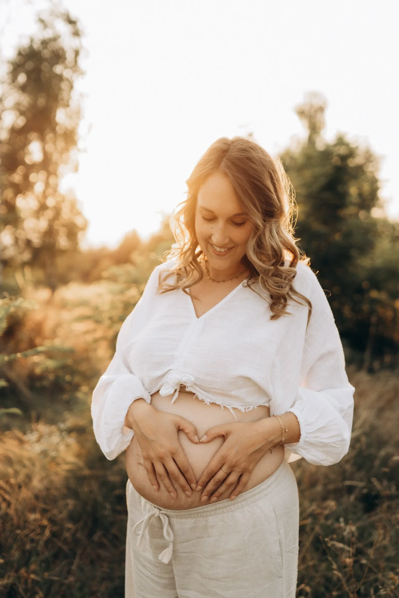 Eine schwangere Frau, lächelnd, im weißen Outfit, macht eine Herzform mit ihren Händen über ihrem Babybauch im Außenbereich bei Sonnenuntergang.