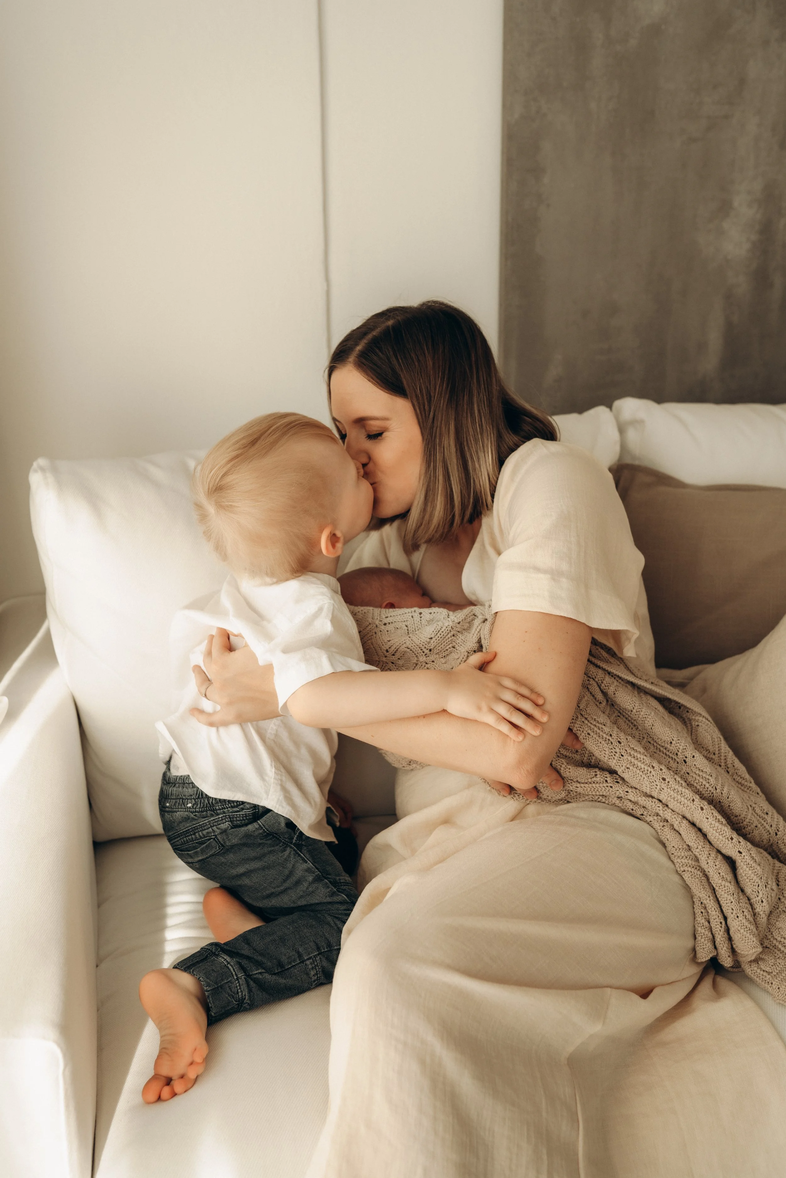 Eine Frau mit braunen Haaren küsst ein Kind und ein Baby im Arm auf einer Couch.