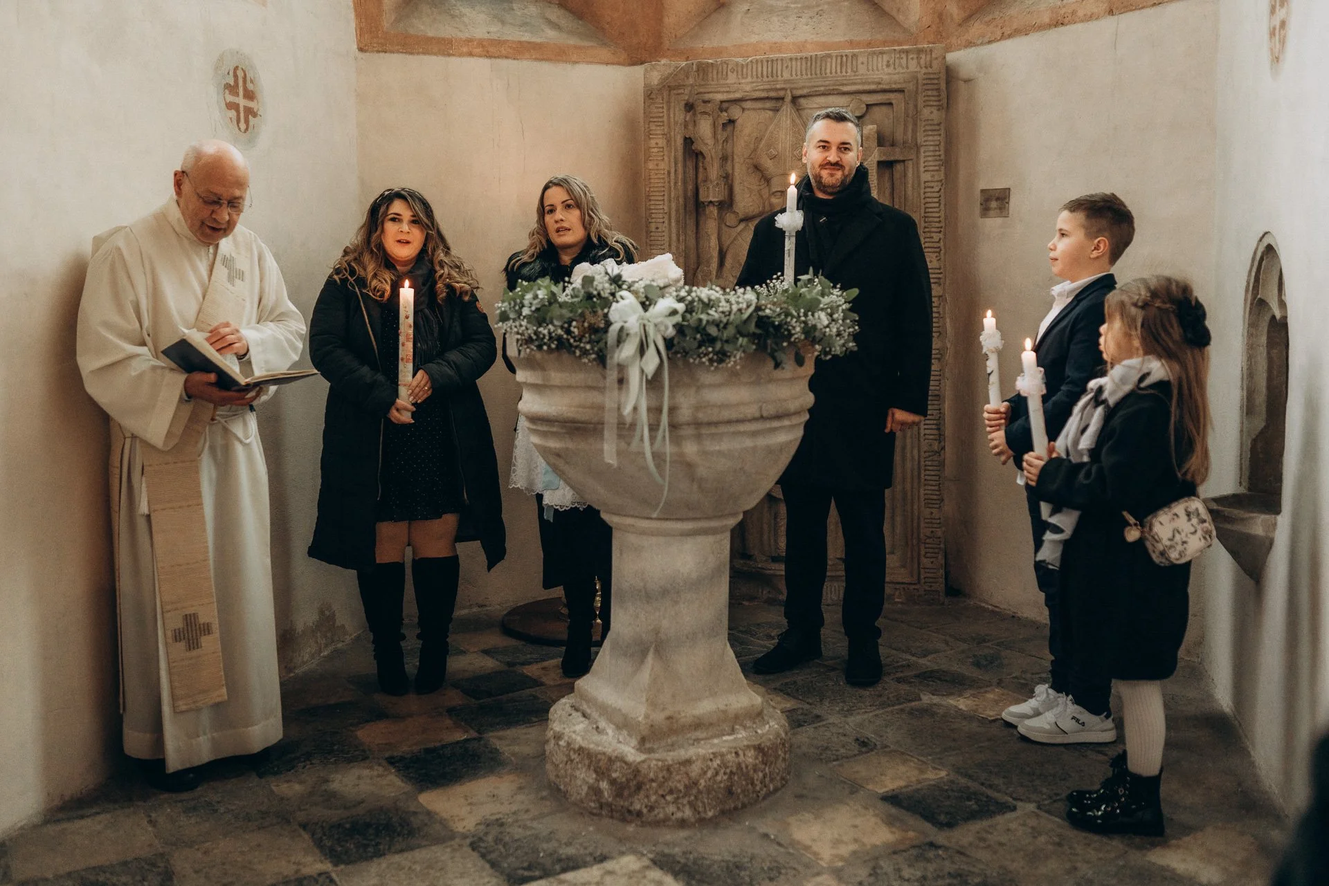 Eine Gruppe von Menschen, darunter ein Priester, versammelt sich in einer Kirche um ein Taufbecken mit Blumen. Sie hält Kerzen und scheint an einer Taufzeremonie teilzunehmen.
