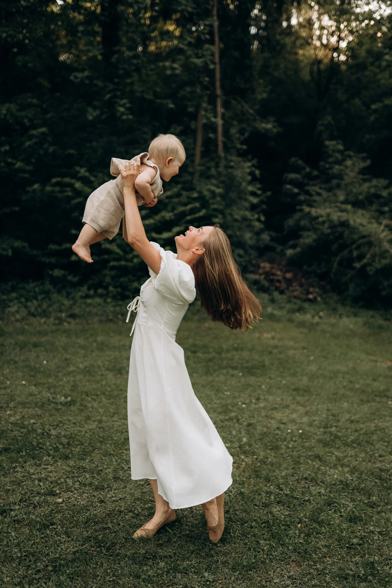 Frau hebt ein lächelndes Baby in die Luft, während sie im Freien auf dem Rasen stehen, umgeben von Bäumen.