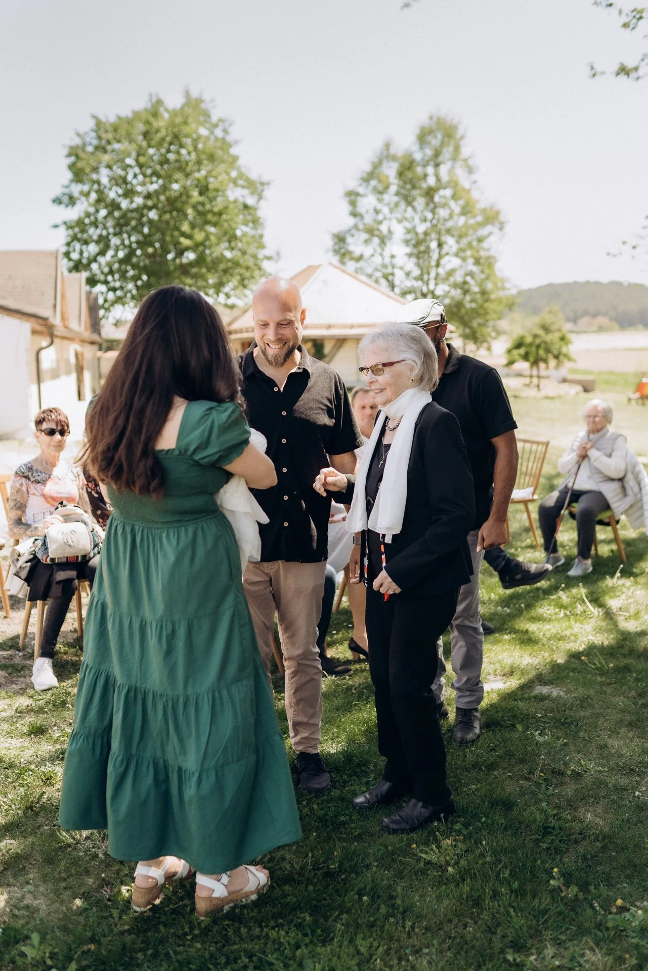 Menschen bei einem Outdoor-Treffen, die sich lachen und unterhalten. Im Vordergrund eine Frau in grünem Kleid, im Hintergrund Bäume und eine ländliche Umgebung.