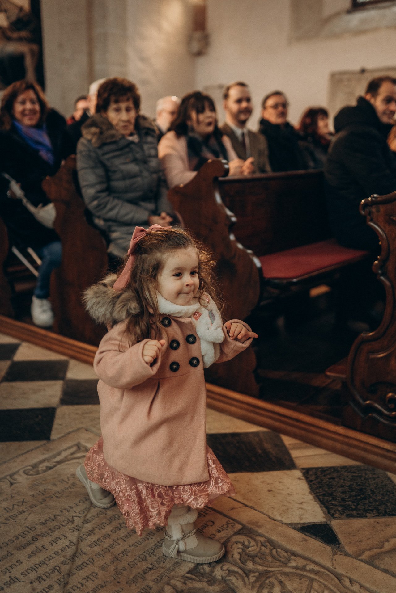 Ein kleines Kind in einem pinkfarbenen Mantel mit Fellkragen und Spitzenrock, das in einer Kirche oder einem ähnlichen Ort steht, während Menschen im Hintergrund sitzen und zuschauen.