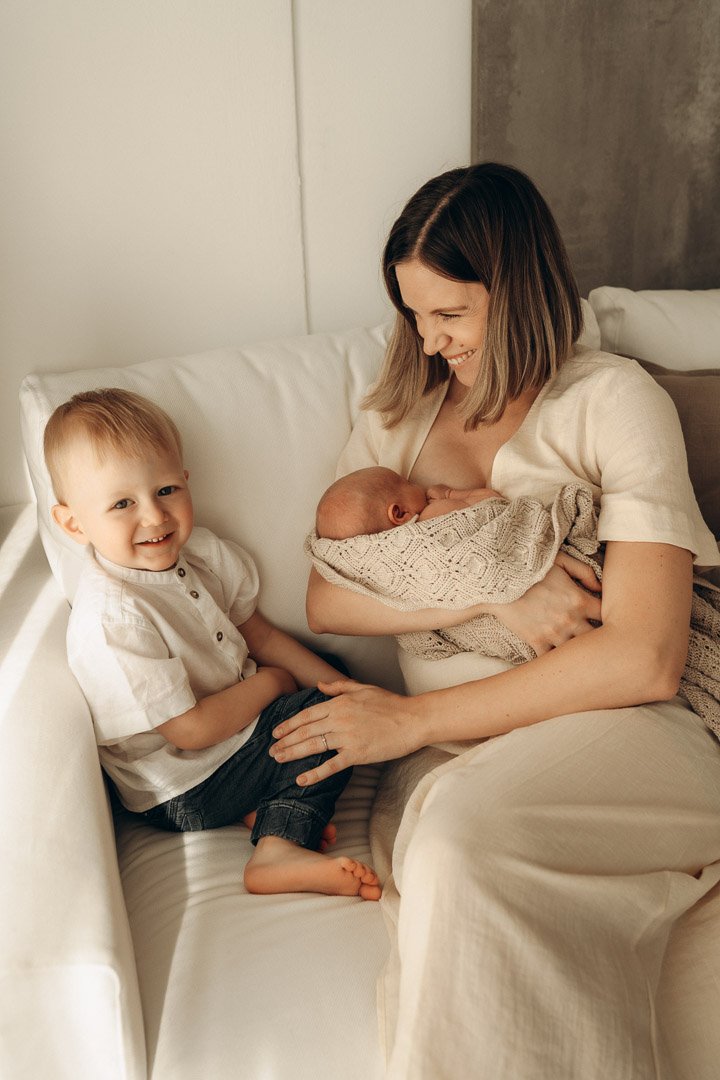 Eine Frau hält ein neugeborenes Baby in den Armen, während ein älterer Junge neben ihr sitzt und lächelt.