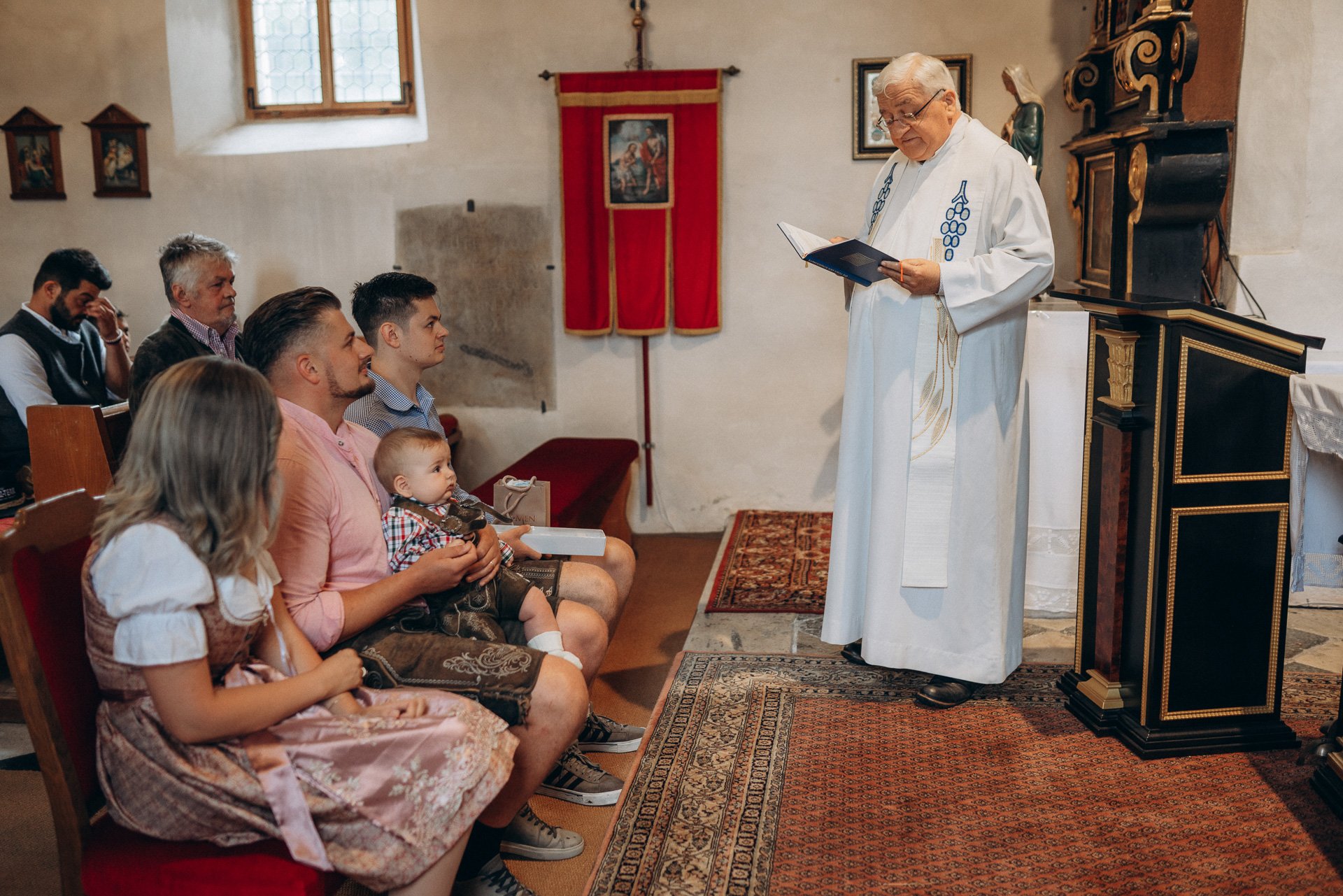 Ein Priester steht in einer kleinen Kirche vor einer Gruppe von Personen, darunter Kinder und Erwachsene, während er einen Gottesdienst oder eine Predigt hält. Die Kirche ist einfach ausgestattet mit religiösen Bildern und einer roten Wand-Flagge im 