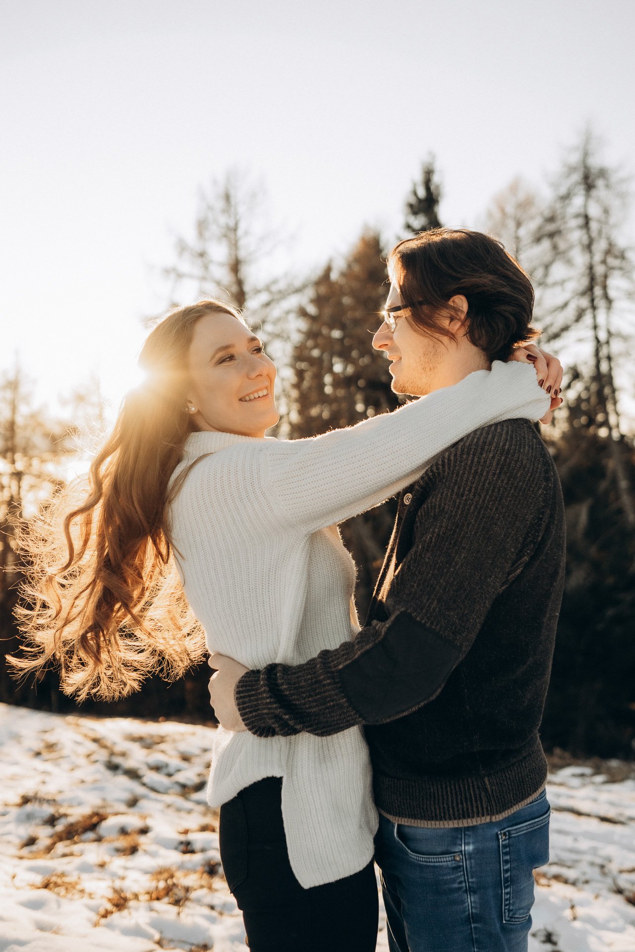 Kuschelige Momente im Schnee: Ein Paar umarmt sich im winterlichen Wald bei Sonnenuntergang und genießt die magische Stimmung während eines Winter-Fotoshootings in Klagenfurt, Kärnten.
