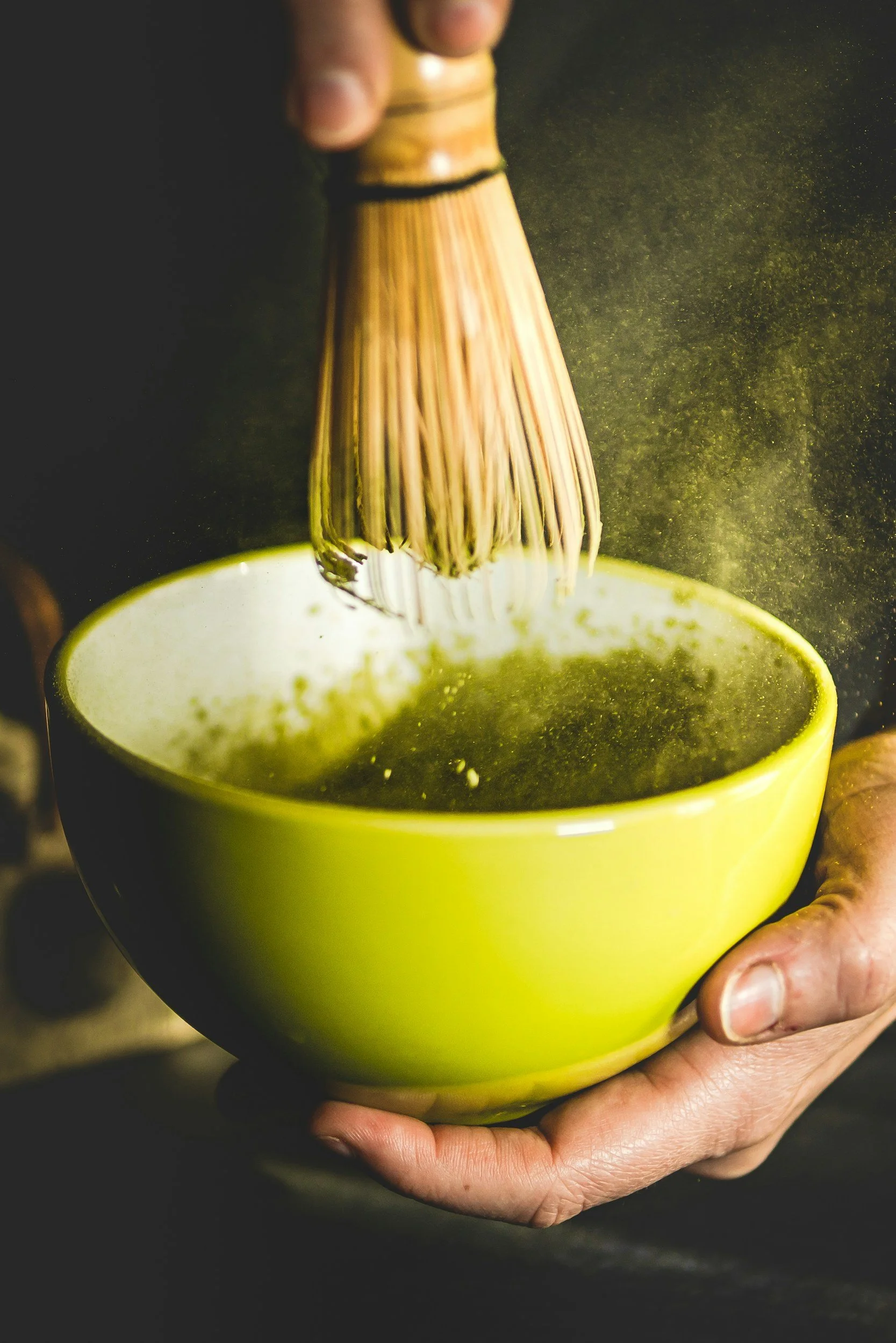 A person holding a green ceramic cup filled with matcha green tea, while whisking the matcha with a bamboo chasen to prepare a traditional Japanese tea.