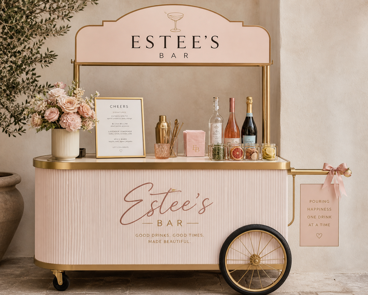 A pink and gold mobile bar cart with the words "Estee's Bar" on the side, decorated with a bouquet of pink roses and other flowers. On top, there are bottles of alcohol, a menu, and glass jars with garnishes. A sign on the right says, "Pouring happiness one drink at a time," and the brand name "Estee's Bar" appears above.