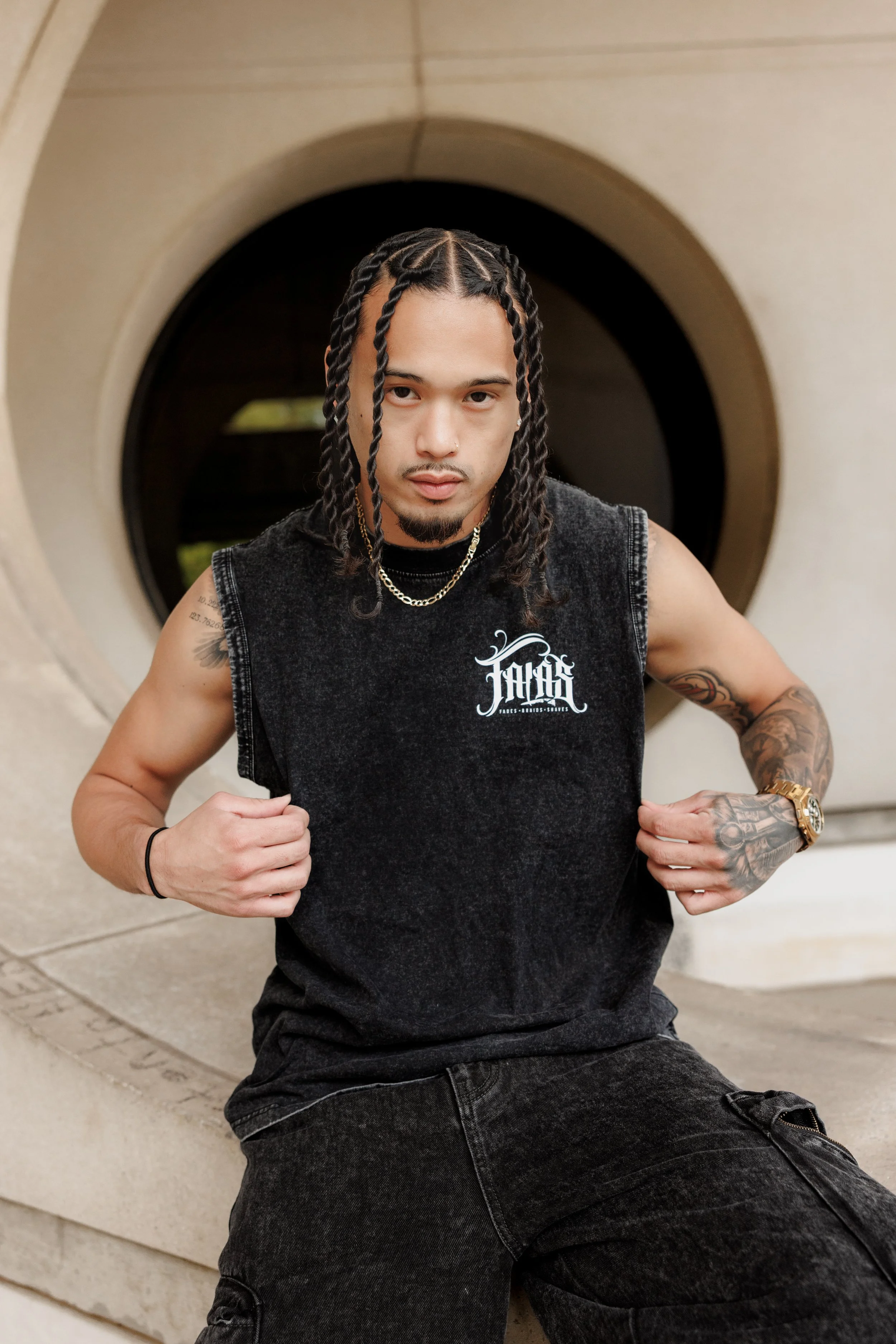 A young man with braided hair, tattoos, and jewelry poses in front of a circular architectural feature.