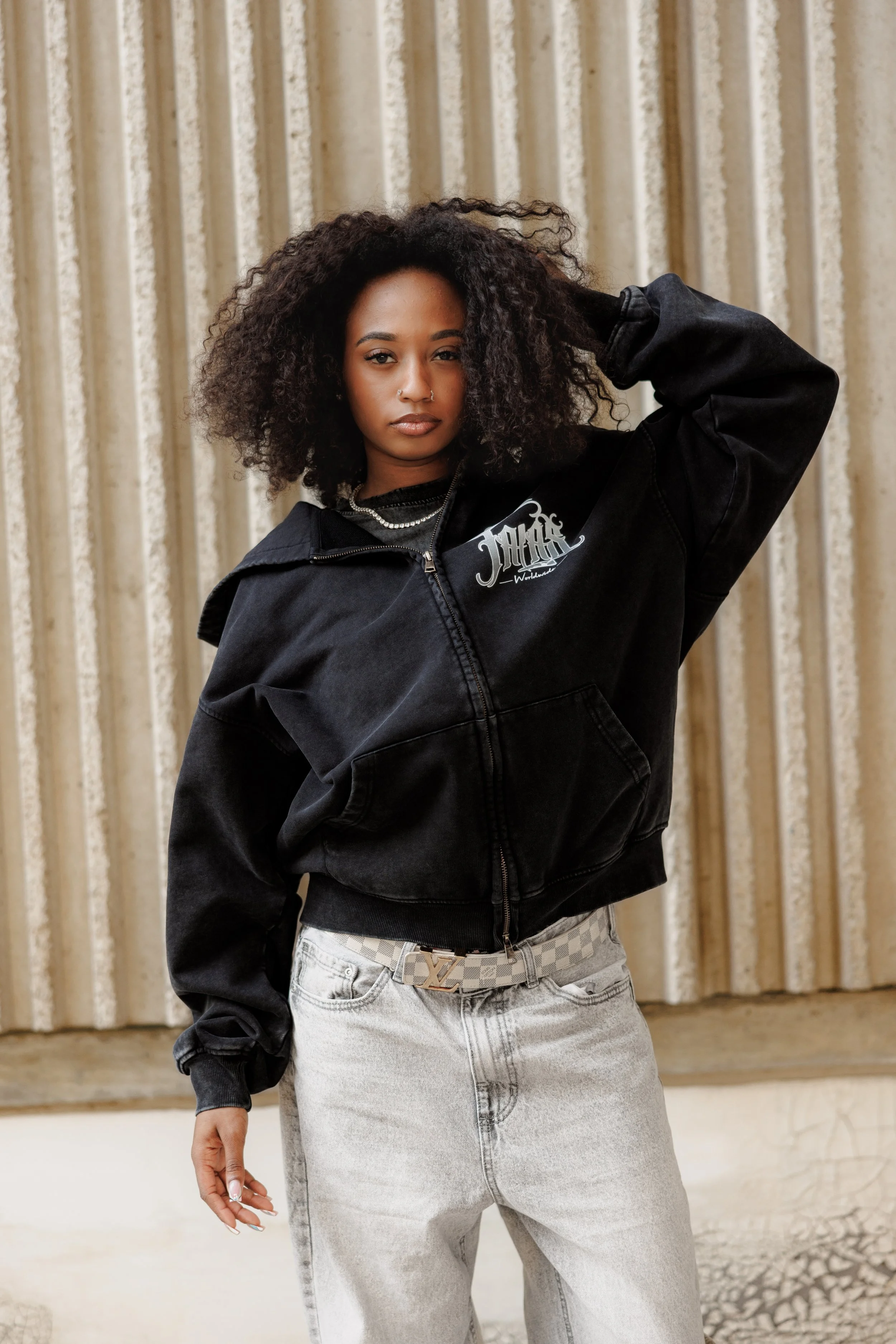 A woman with curly black hair wearing a black jacket with logo, white accessories, and light-colored jeans standing against a beige, textured curtain backdrop.