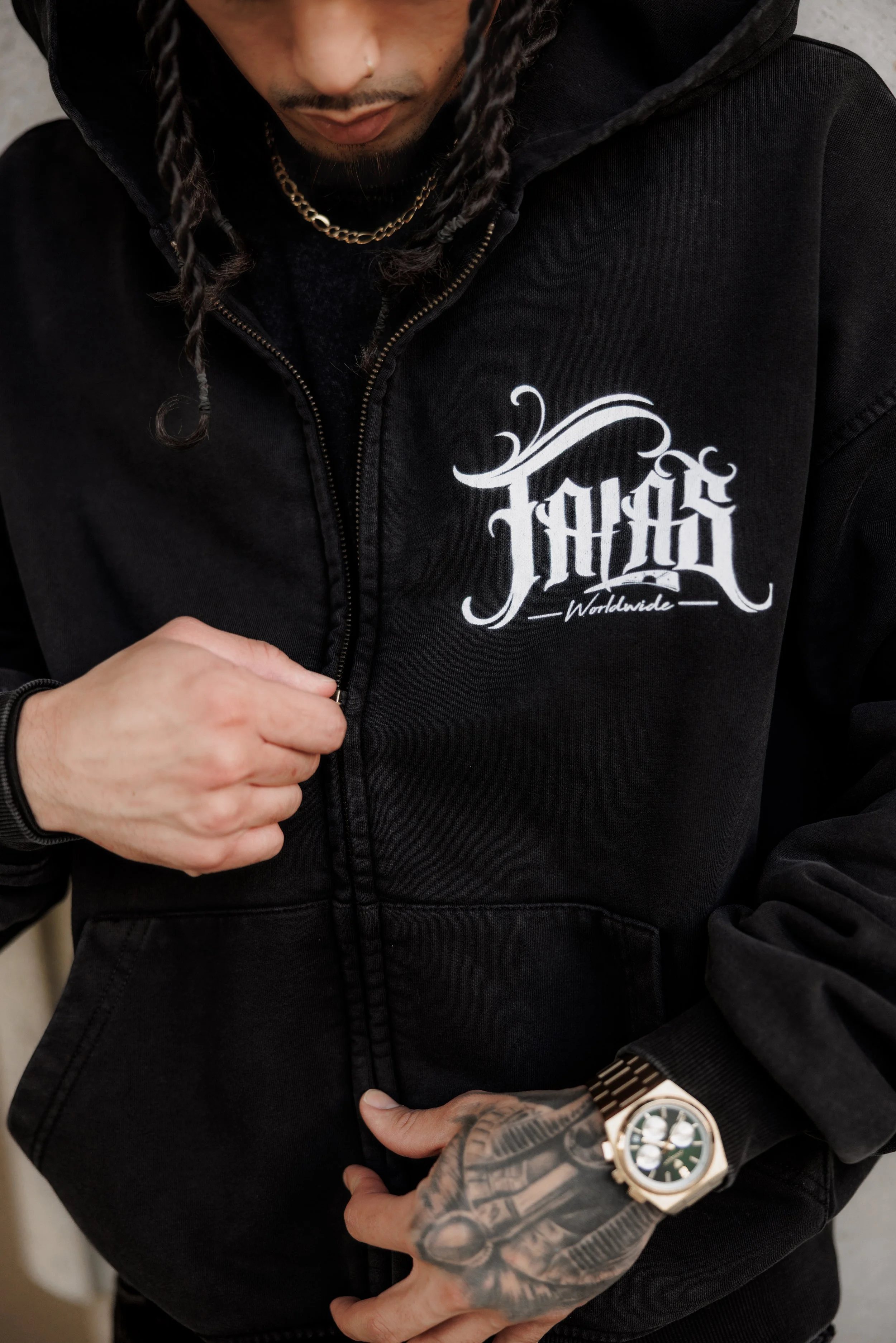 Close-up of a person wearing a black hoodie with white graphic text 'HUSTLE Worldwide' on the chest, gold chain necklace, gold watch, and tattooed left hand adjusting the zipper.