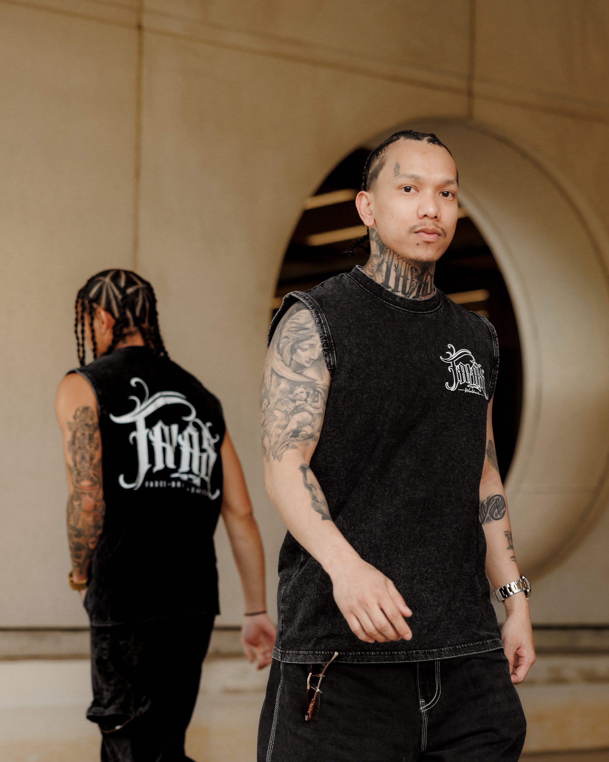 Two men with tattoos, dressed in black sleeveless shirts, standing back-to-back in an indoor setting, with one man facing forward and the other with back turned.