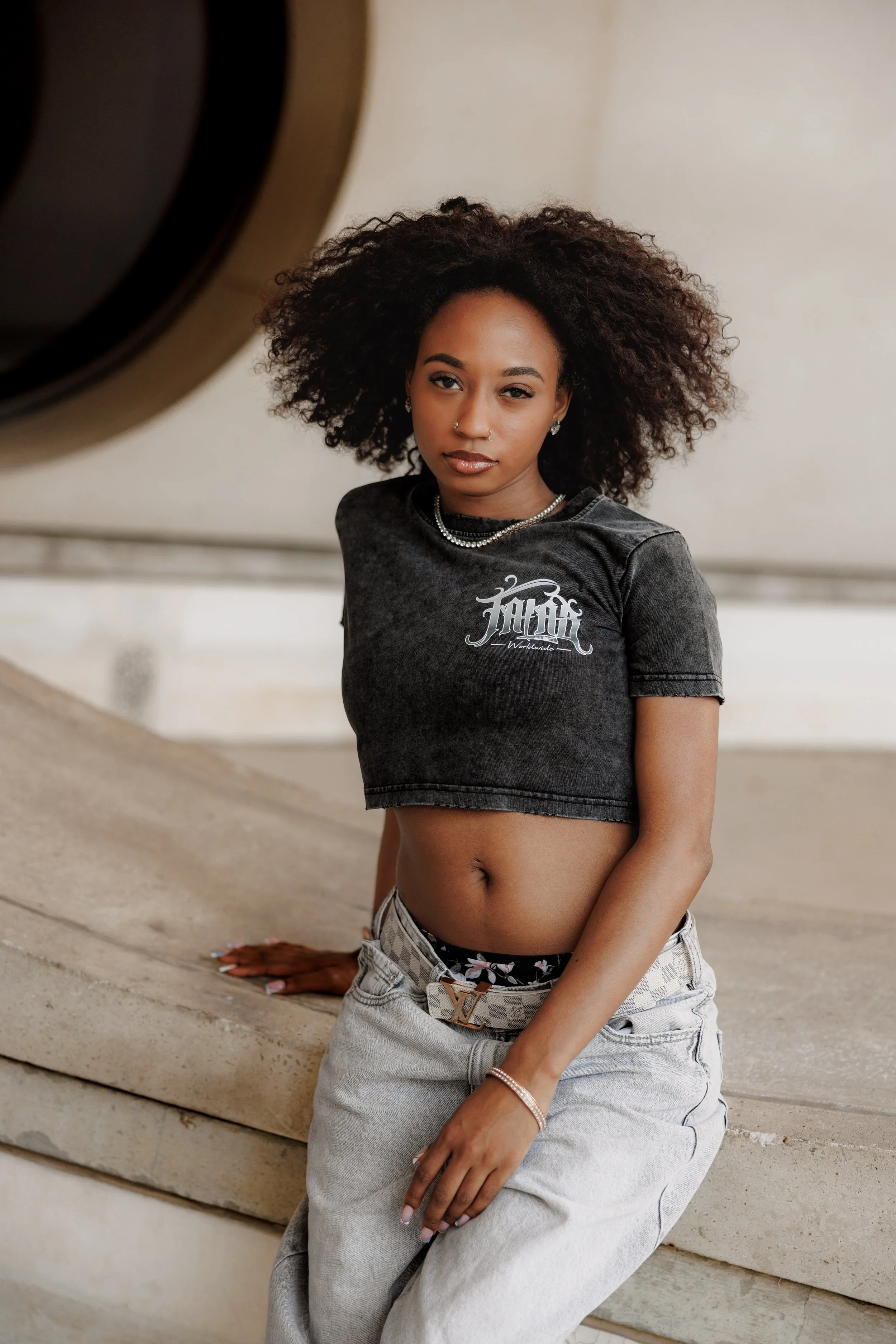 A woman with curly hair wearing a black cropped T-shirt with a graphic and light-colored jeans, sitting on a concrete ledge in an urban setting.