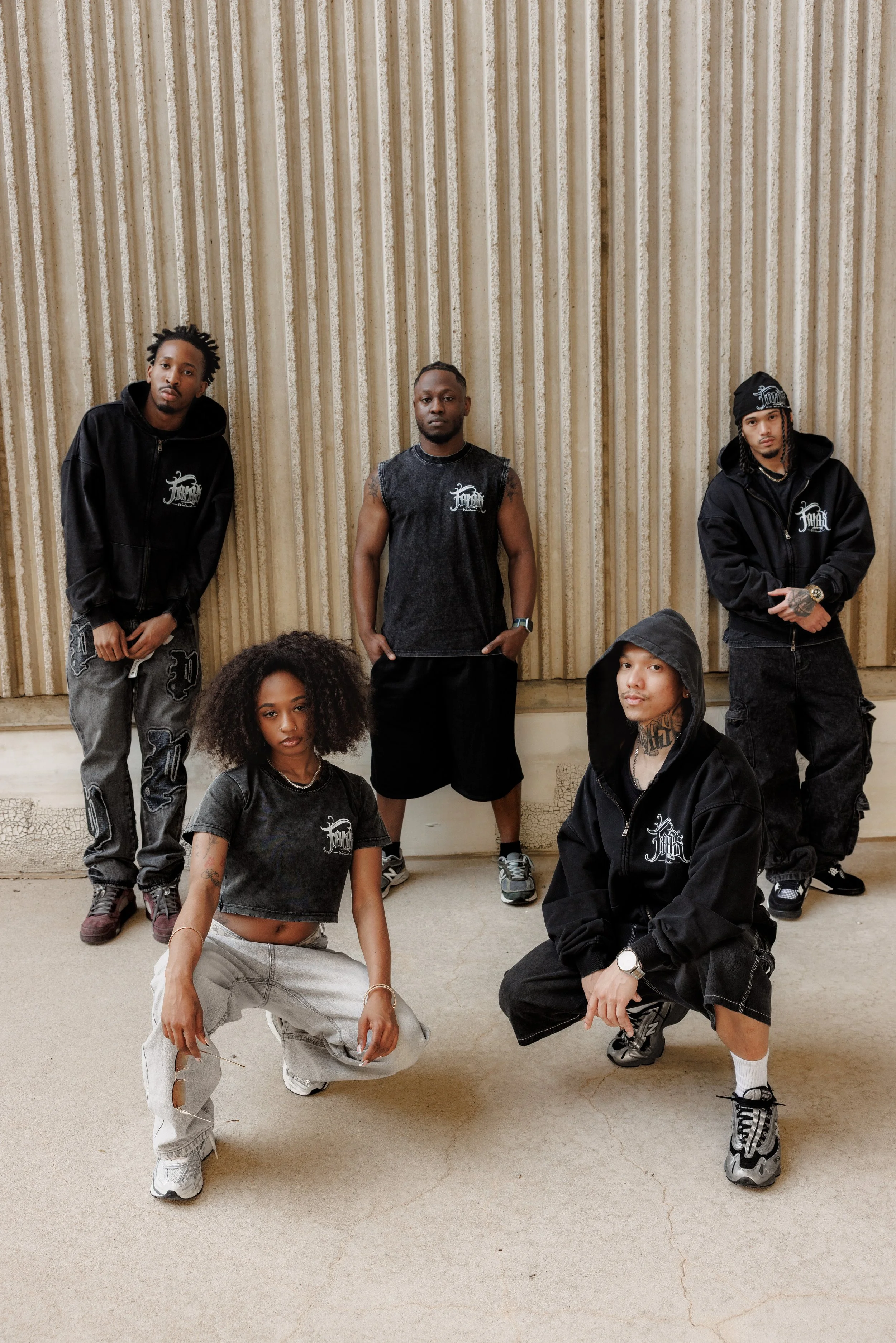 Group of six young adults posing against a corrugated metal wall, some standing and some squatting, dressed in casual streetwear, with various hairstyles and accessories.